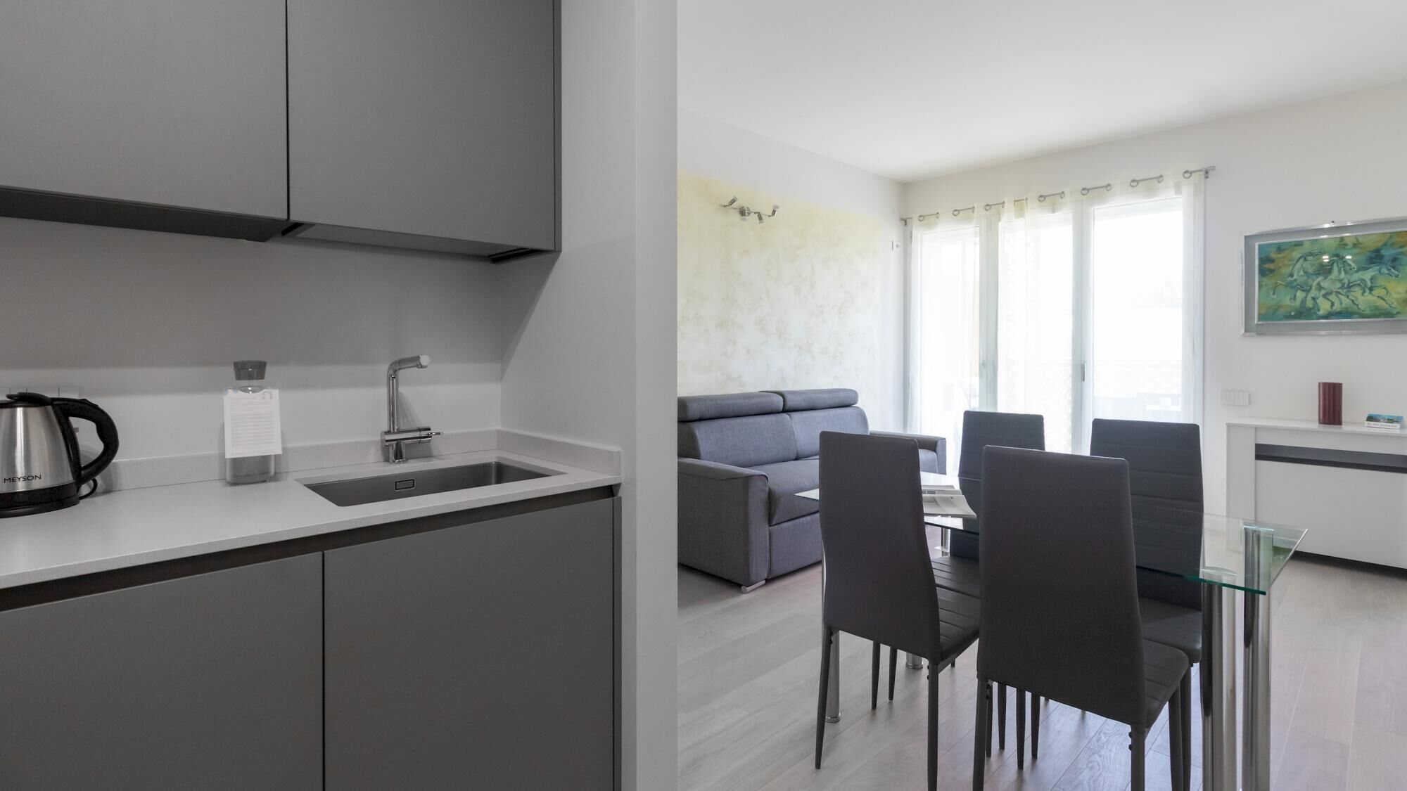 A modern kitchen with a sink, kettle, and cabinets. There is a dining area with a table and chairs. A gray sofa and a window with curtains are visible in the background.