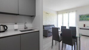 Una cocina moderna con un lavamanos, hervidor y armarios. Se ve un comedor con una mesa y sillas. En el fondo, un sofá gris y una ventana con cortinas.