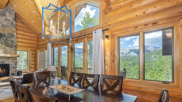 Un intérieur en bois comprenant une table à manger, des chaises, une cheminée et de grandes fenêtres offrant une vue sur les montagnes.