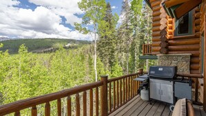 Une terrasse en bois avec une cheminée en pierre et un barbecue, donnant sur une vallée boisée.