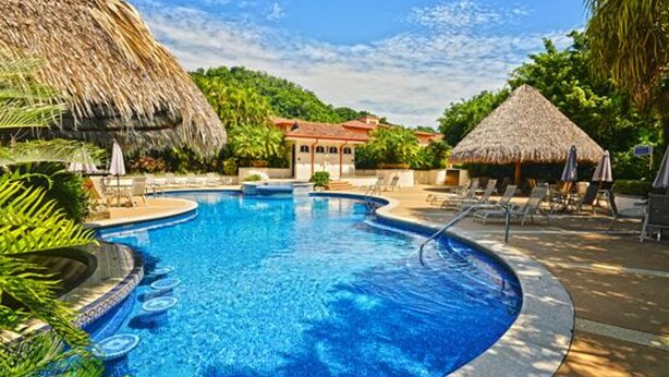 A swimming pool with a thatched roof structure, surrounded by lounge chairs and umbrellas, with a building in the background. - Colina 9E 2 bdr 2 Bath Family (Jacó)