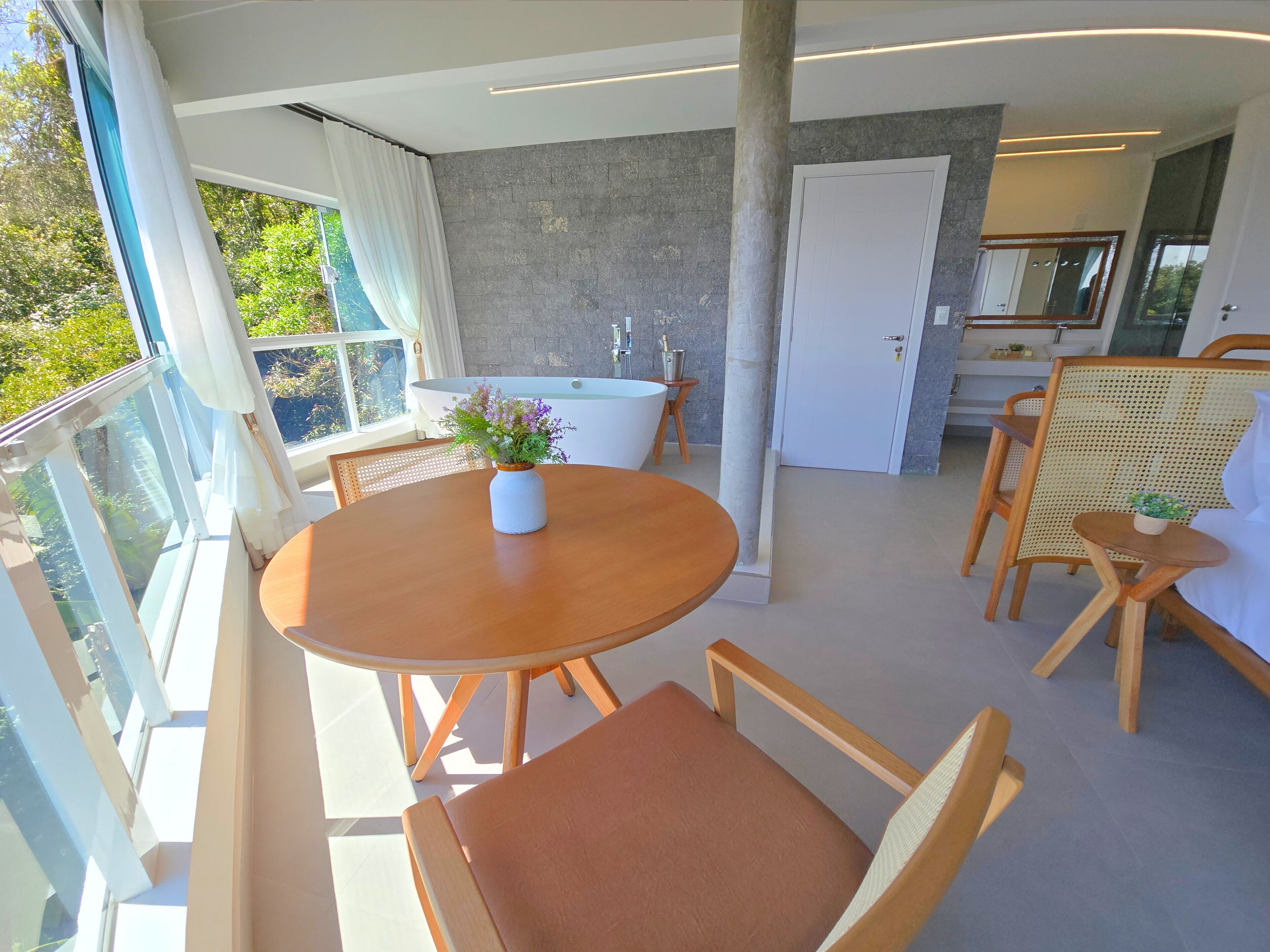 A modern, well-lit room with a round wooden table, two chairs, a small potted plant, a bathtub, and a large mirror.