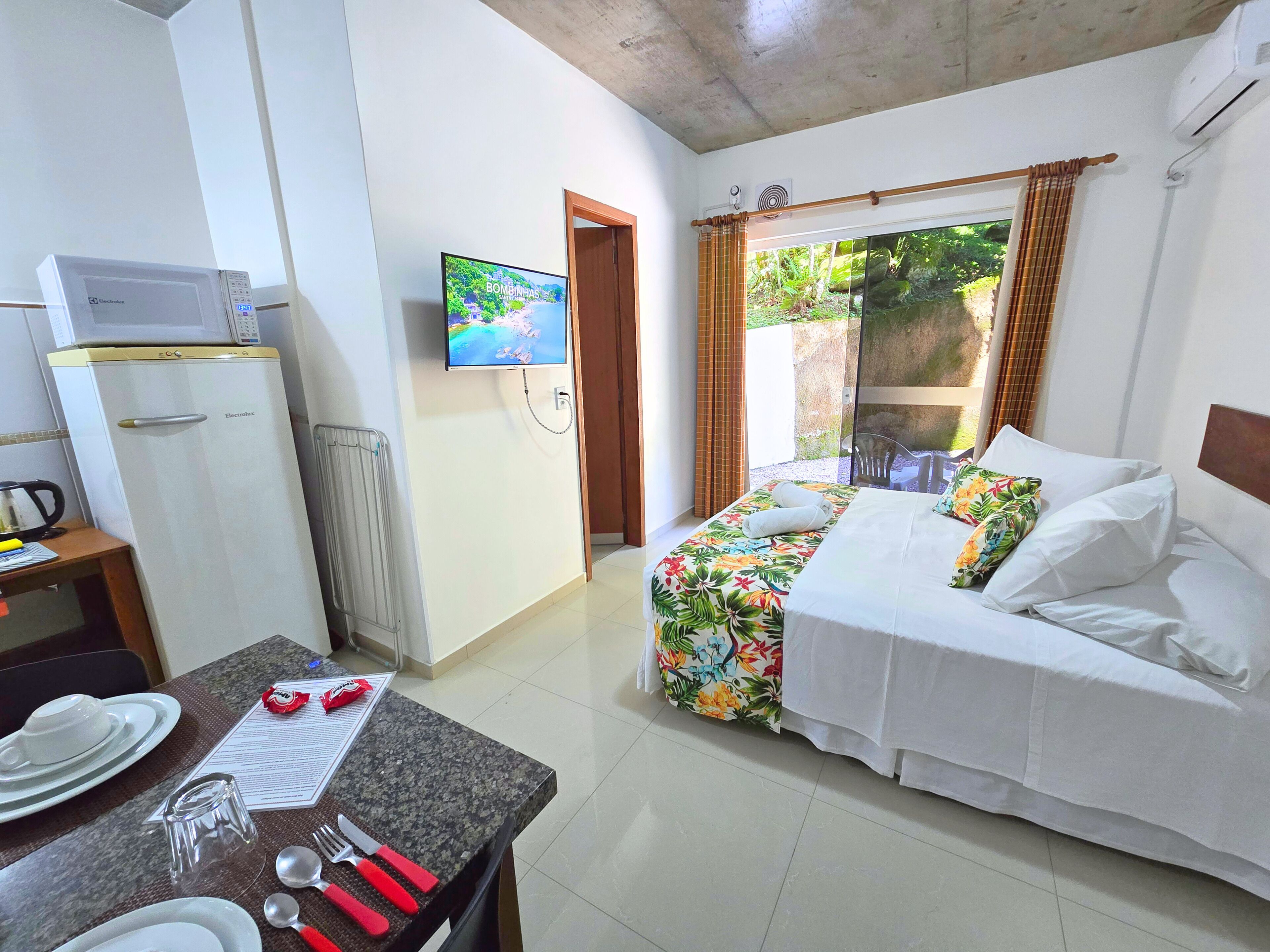 A hotel room with a bed, a television, a microwave, a dining table with cutlery, and a window with curtains.