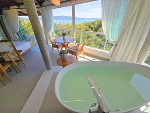 A room with a large bathtub, a dining table with chairs, and a view of the sea and hills. - Refúgio da Lagoinha (Bombinhas)