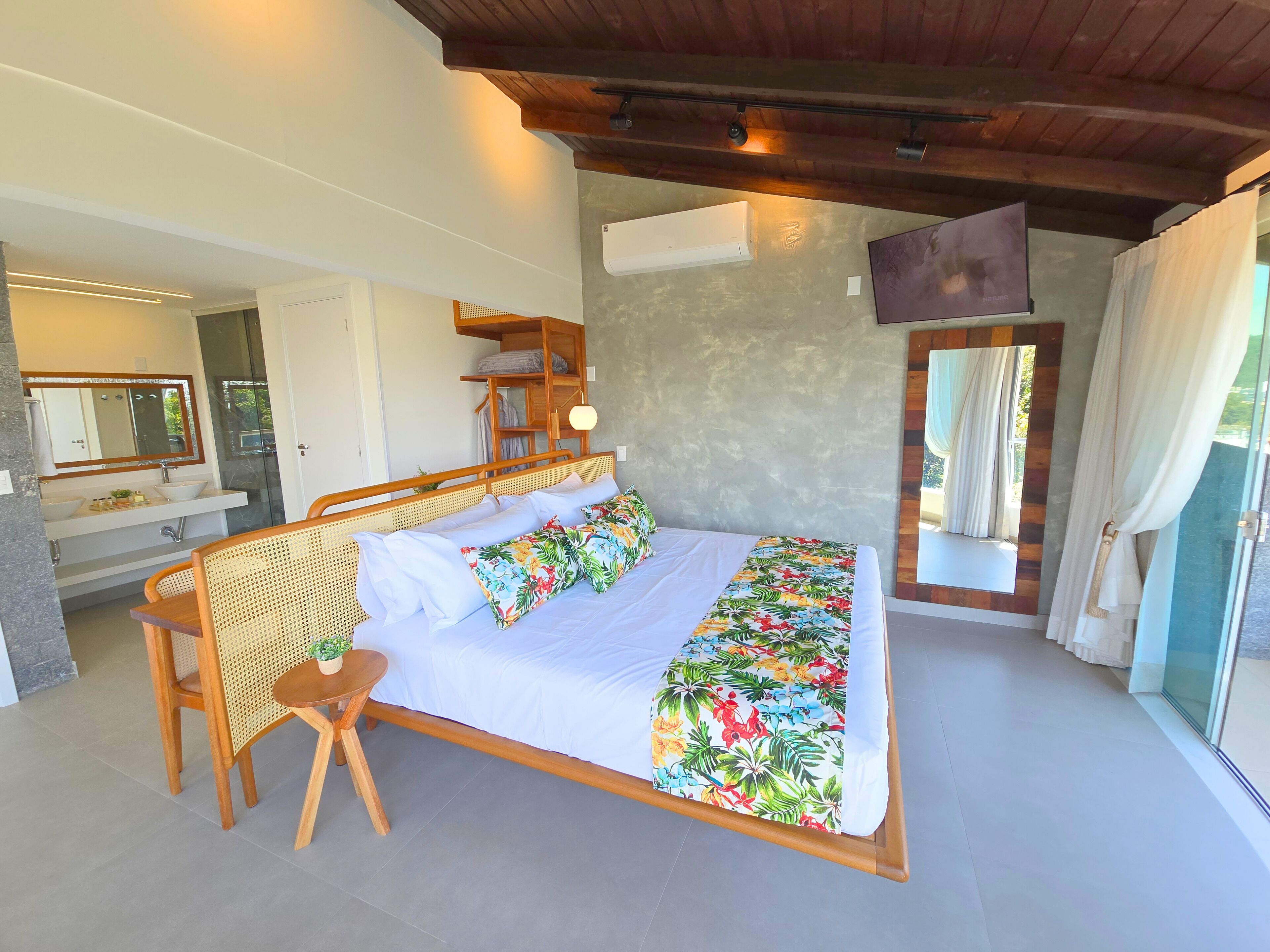 Une chambre à coucher avec un lit, une tête de lit en bois, une table de chevet avec une plante, un miroir, et une salle de bain avec un lavabo et un miroir.