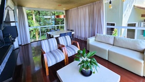 Una sala de estar moderna con sofá, sillas, una mesa de centro y una zona de comedor con mesa y sillas. Hay un ventanal con cortinas, un techo con vigas a la vista y lámparas colgantes.