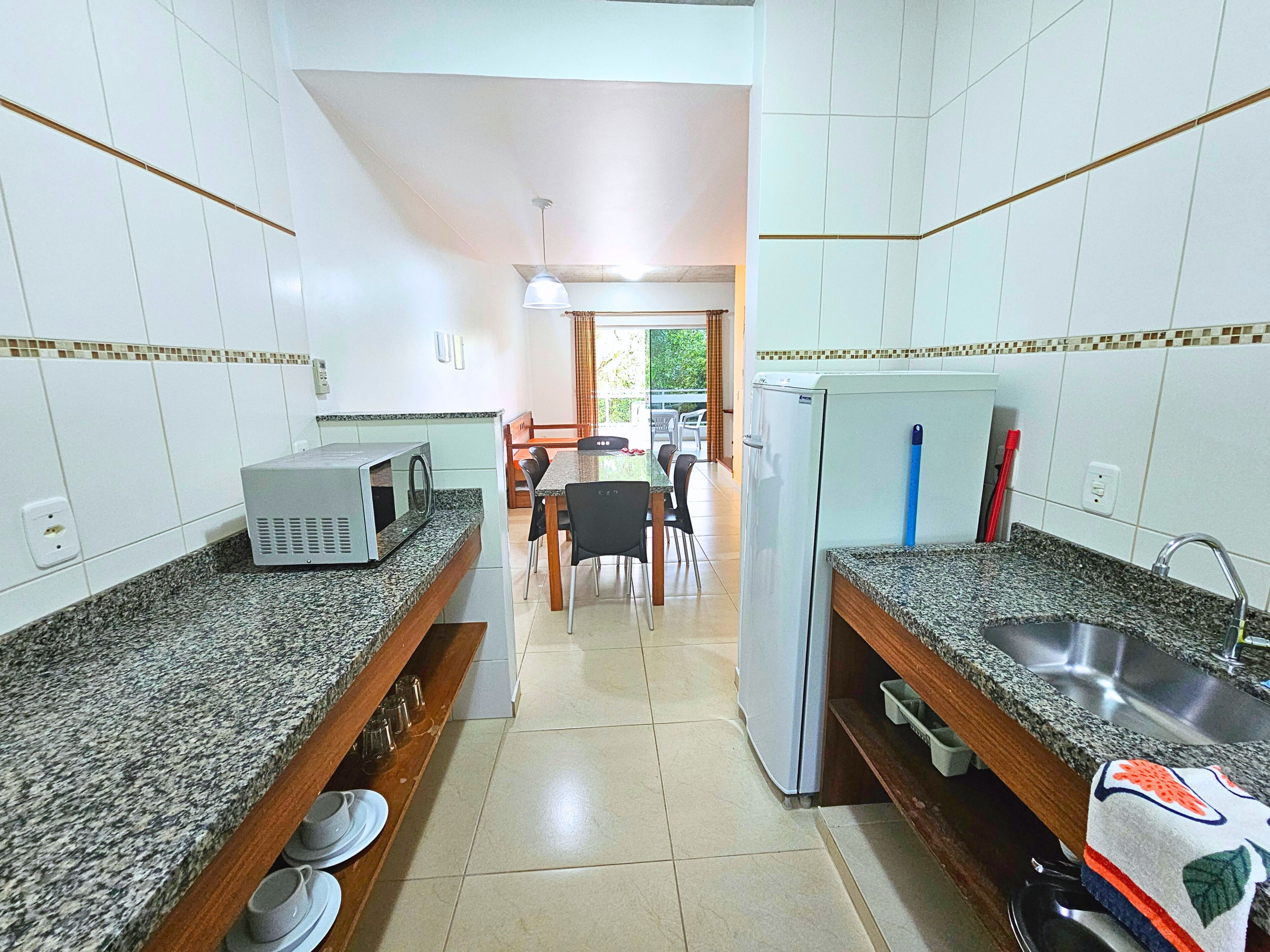 A compact kitchen with granite countertops, a microwave, a refrigerator, and a sink. There is a dining area with chairs and a table in the background.