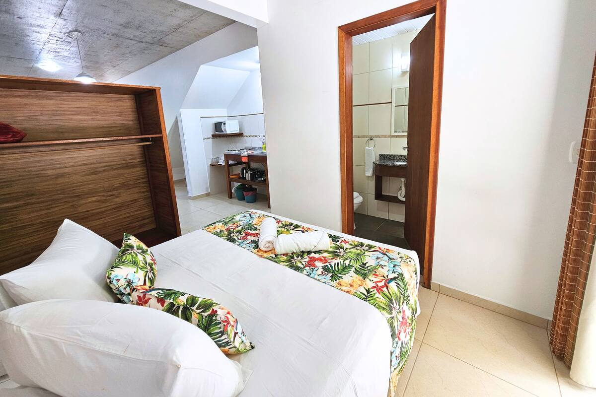 Quarto com cama, cabeceira de madeira, mesa de cabeceira e vista para uma cozinha e banheiro através de uma porta aberta.