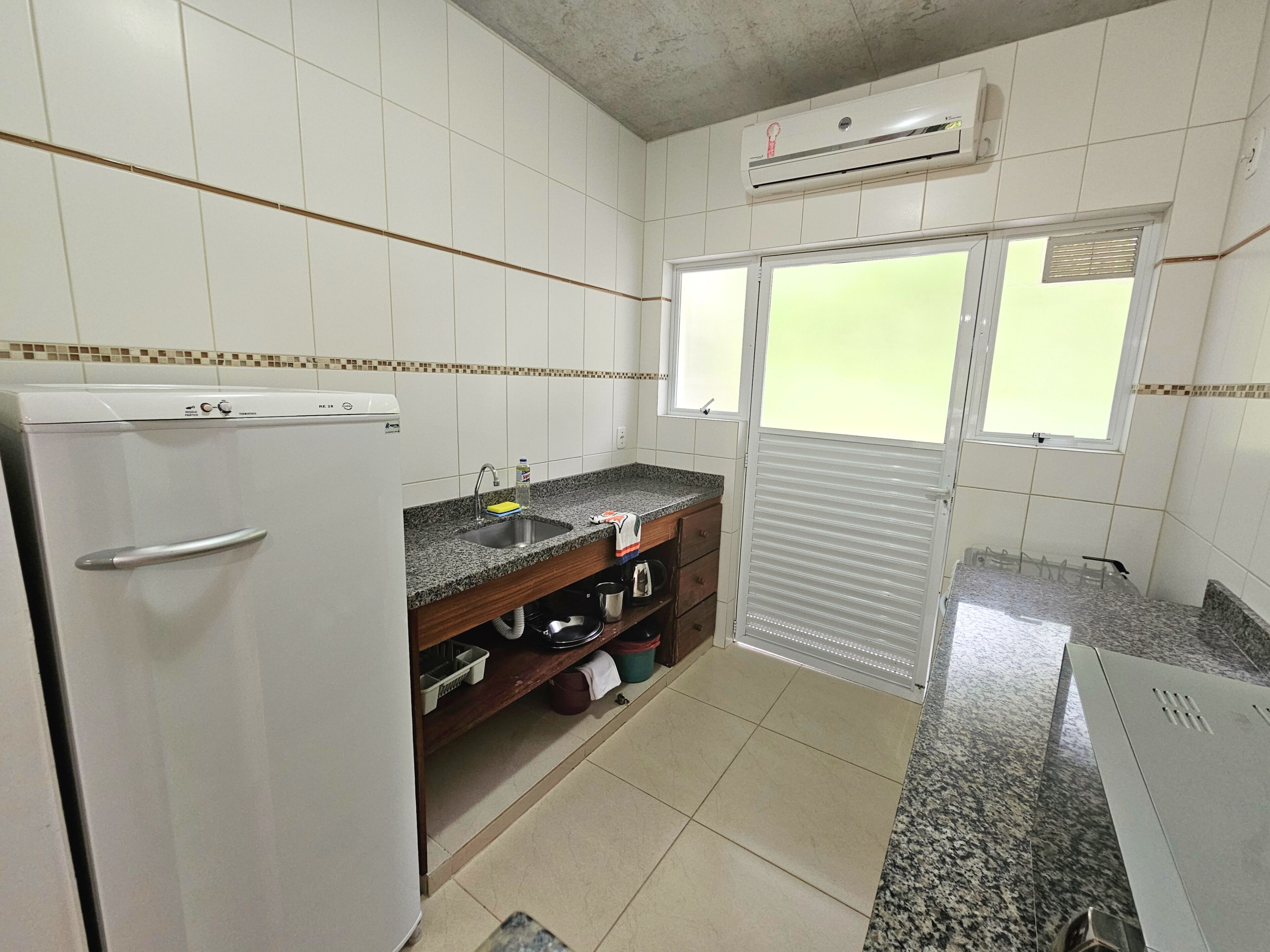 Uma cozinha compacta com paredes revestidas de azulejos brancos, frigorífico embutido, pia e janela com persianas.