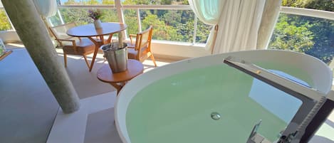 A modern bathroom with a large bathtub, a round table with chairs, and a view of the sea and mountains.