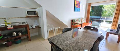 Cocina con microondas, lavamanos y diversos utensilios. Hay un comedor con mesa y sillas. En la pared hay un cuadro, y se ve una ventana con cortinas.