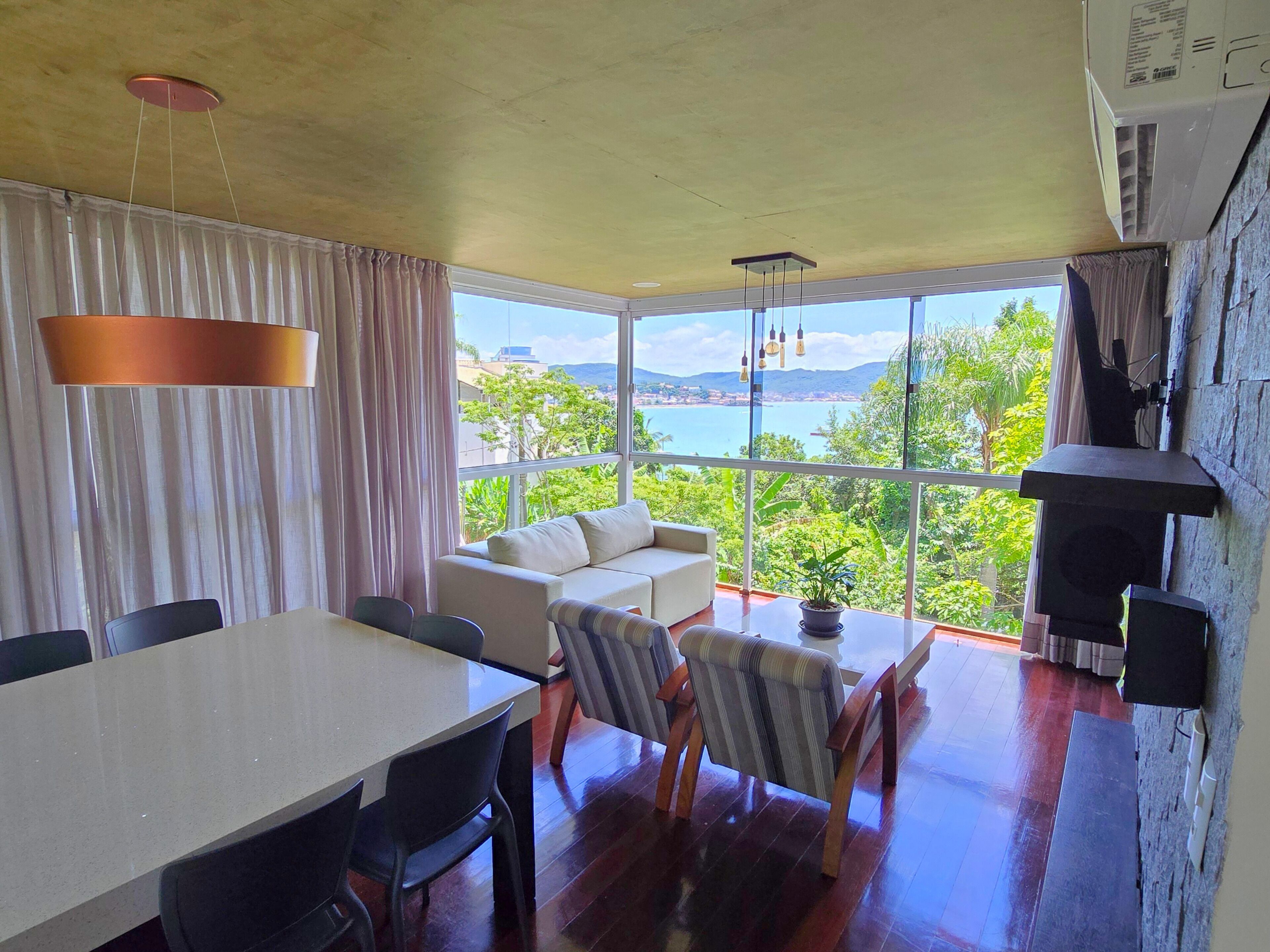 Un comedor moderno con una mesa grande y sillas, un sofá con cojines, y vistas al mar y vegetación a través de amplias ventanas.