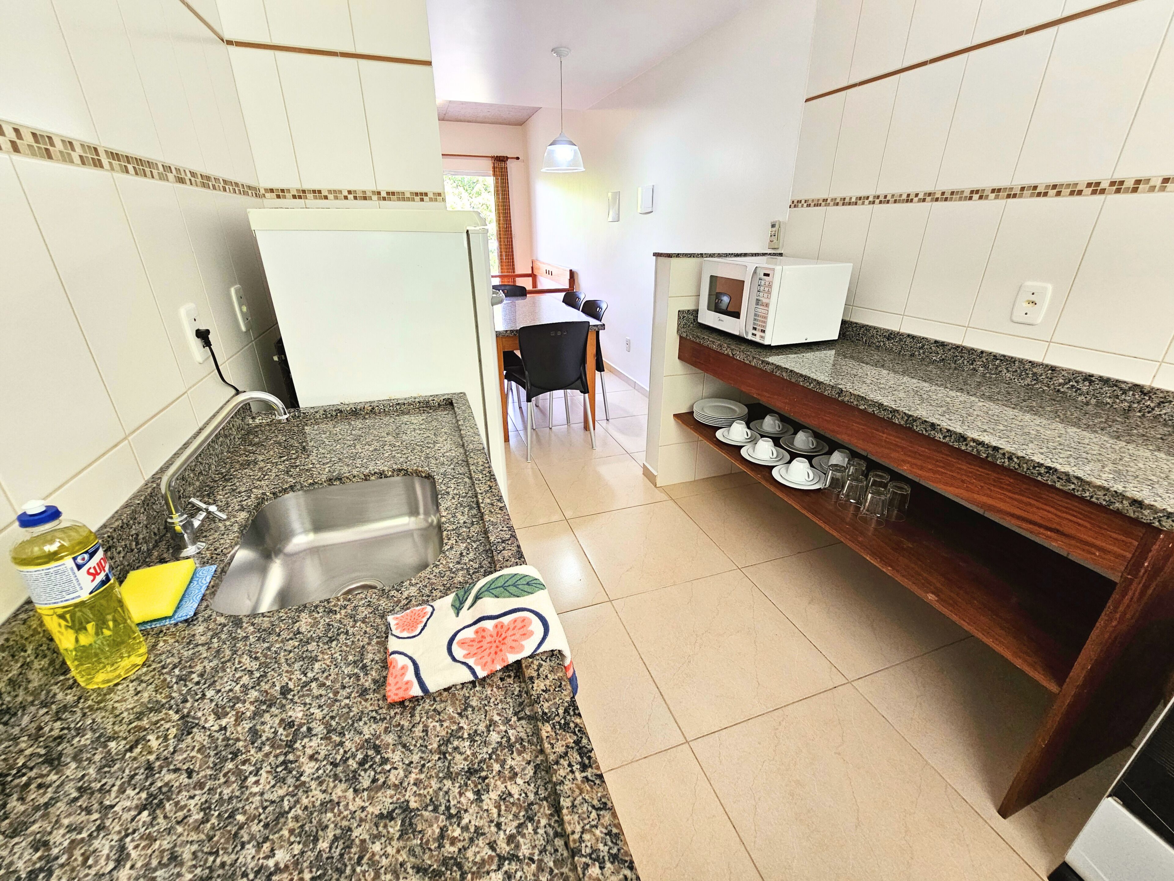 A compact kitchen with a granite countertop, stainless steel sink, and a microwave. There is a dining area visible in the background.