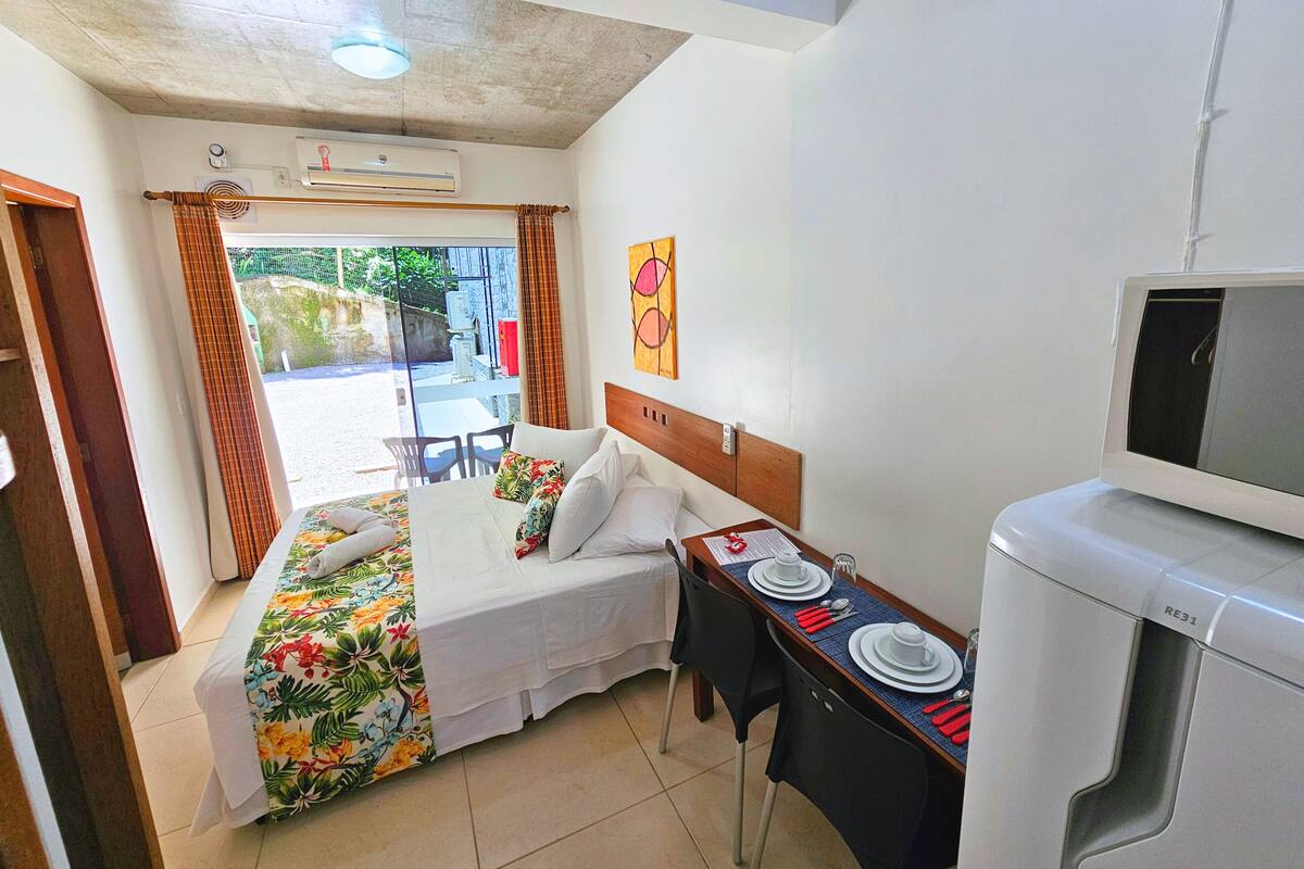 Quarto de hotel com cama, secretária com cadeira, televisão e mesa de jantar posta para dois.