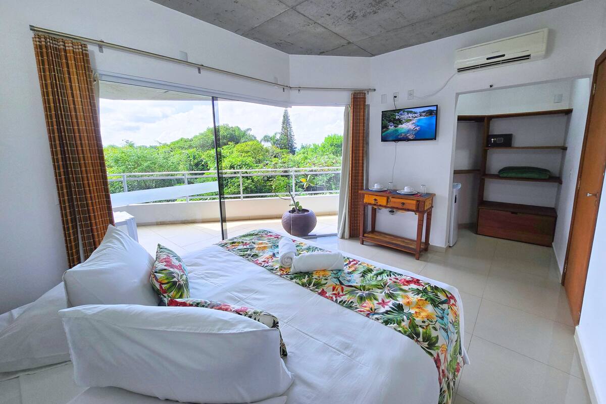 Quarto de hotel com cama, televisão, mesa pequena e varanda com vista para área verde.