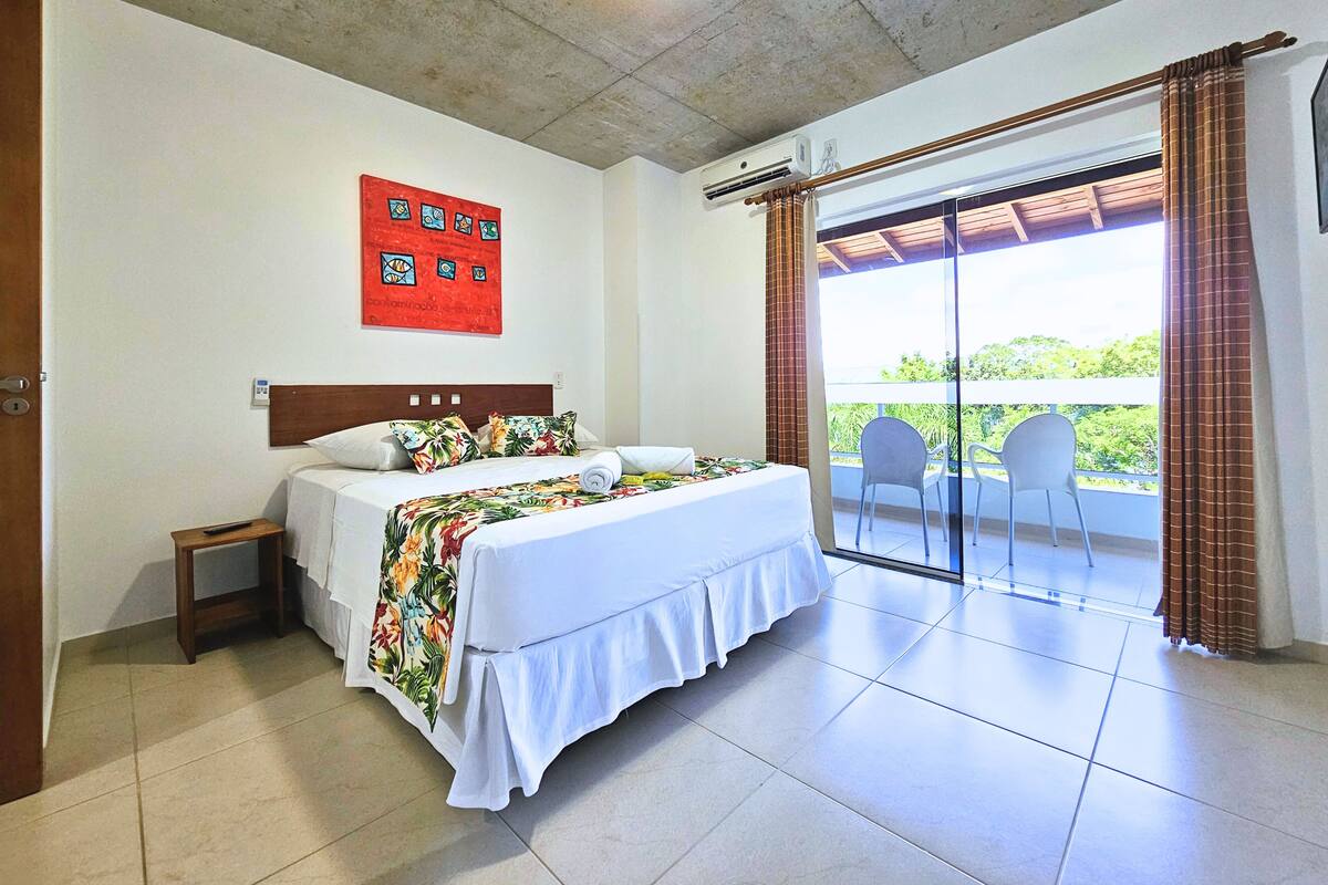 Quarto de hotel com cama, mesa de cabeceira, janela com cortinas e vista para o verde lá fora.