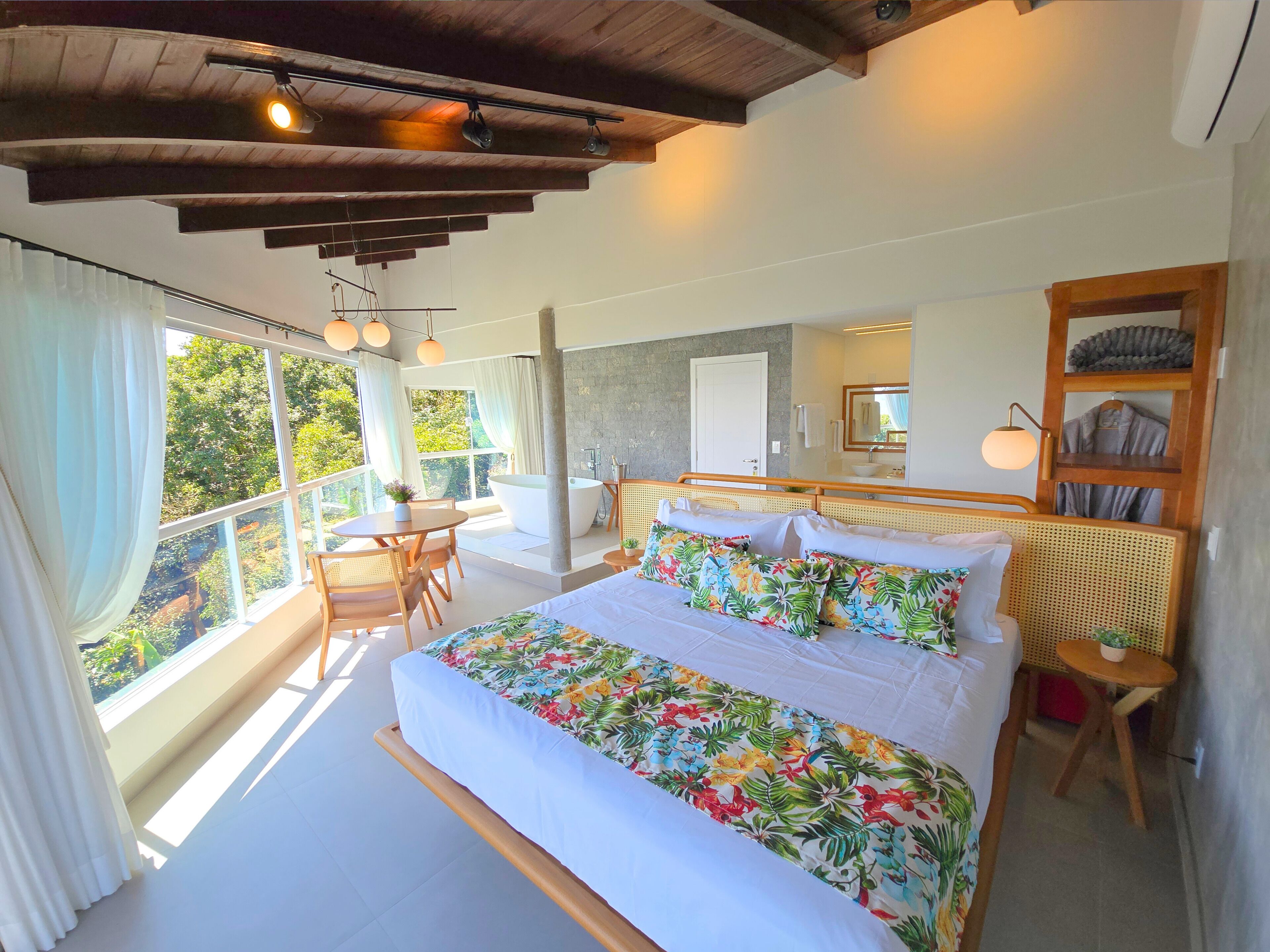 A spacious bedroom with a large bed, a sitting area with a table and chairs, a bathtub, and a view of greenery outside.