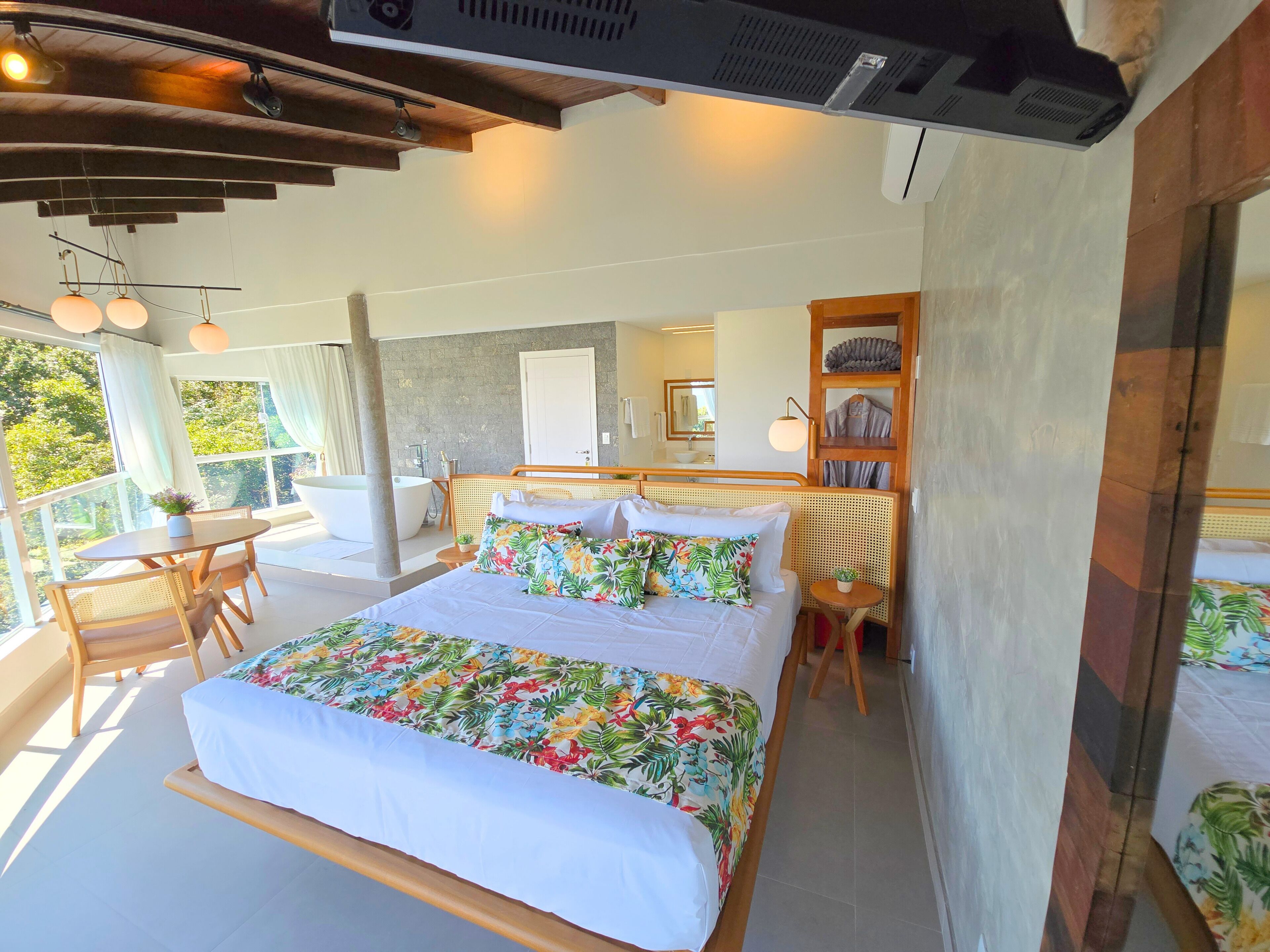 A bedroom with a large bed, a sitting area with a table and chairs, a bathtub, and a view of greenery outside.