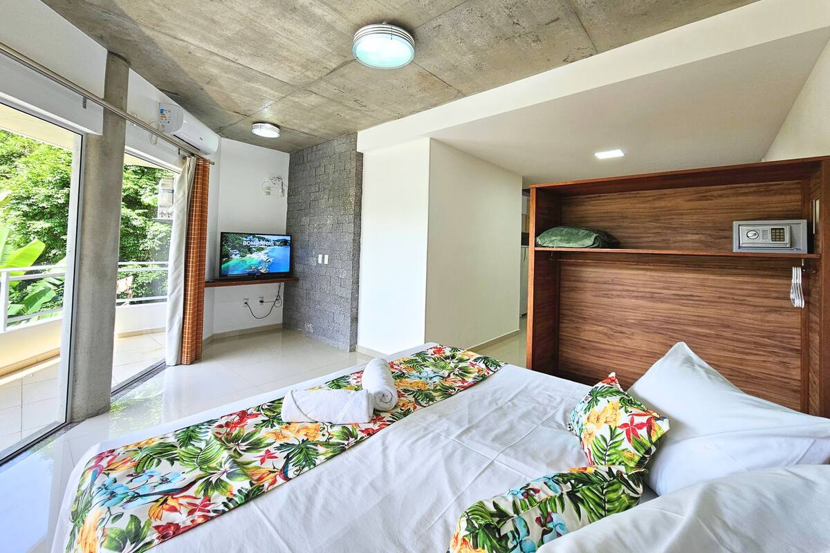 Quarto de hotel com uma cama grande, televisão na parede e vista para o verde lá fora.