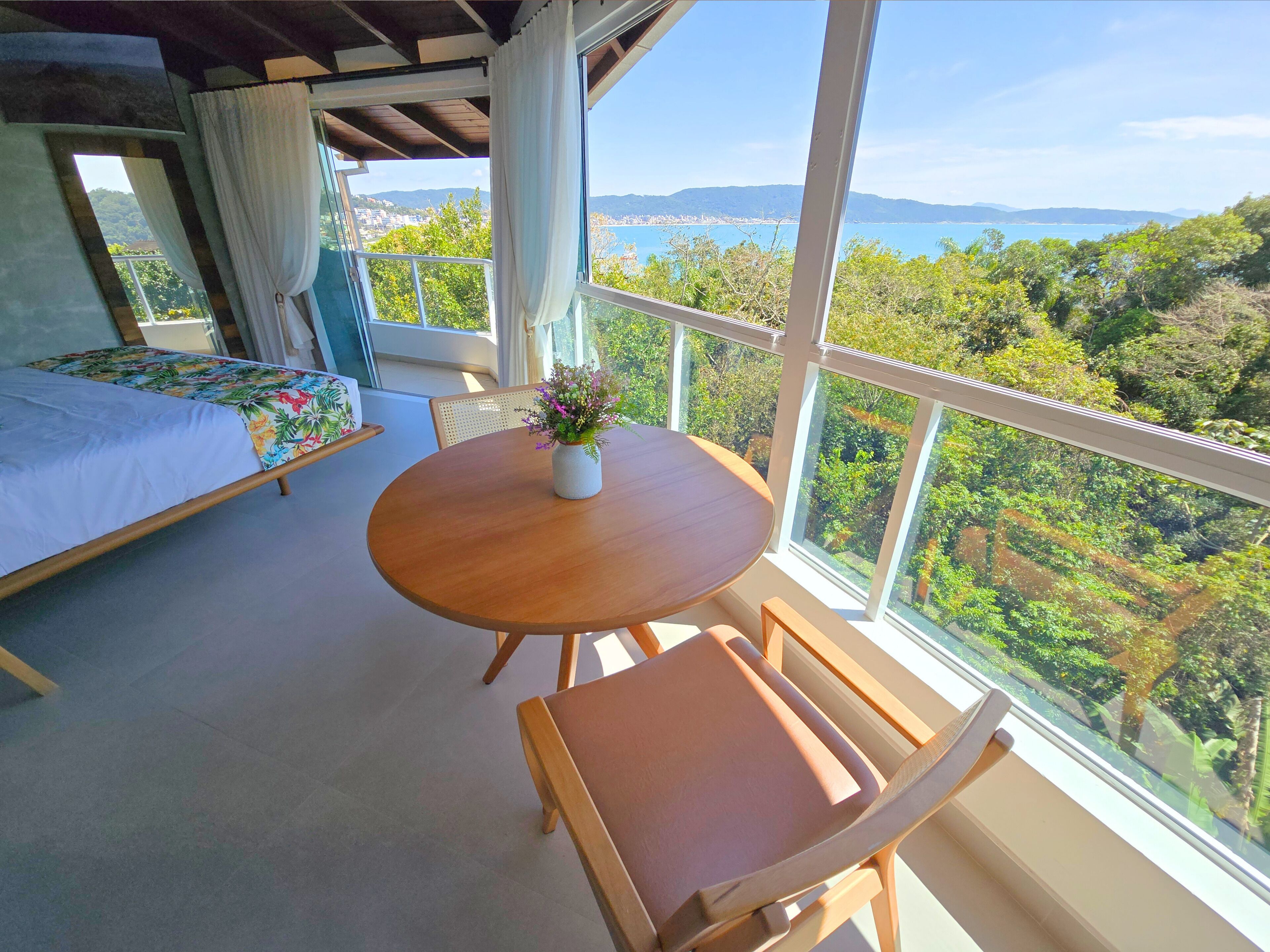A bedroom with a bed, a round wooden table with a vase, and a view of a coastal landscape.