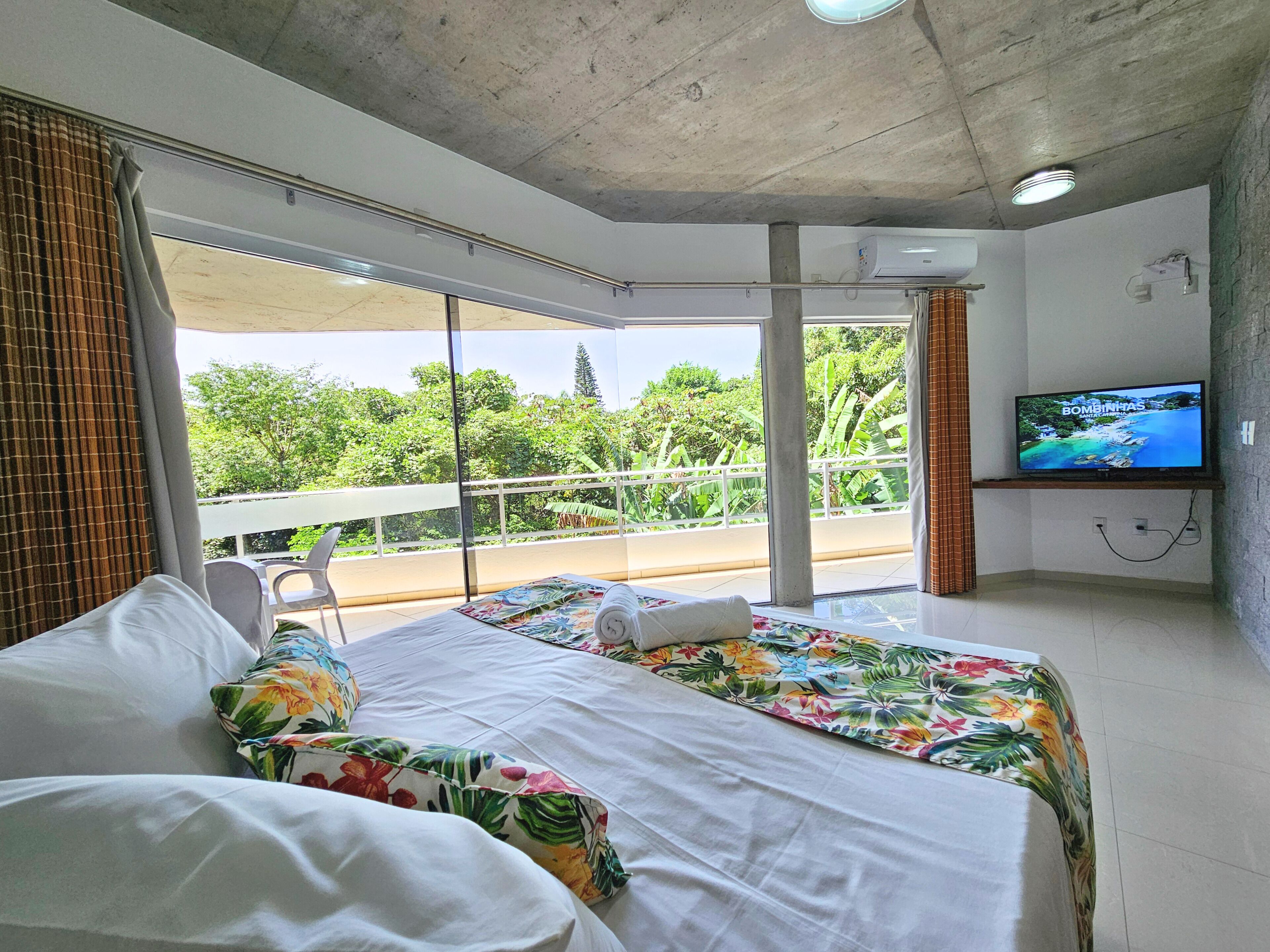 A hotel room with a large bed, a TV, and a balcony with a view of greenery.