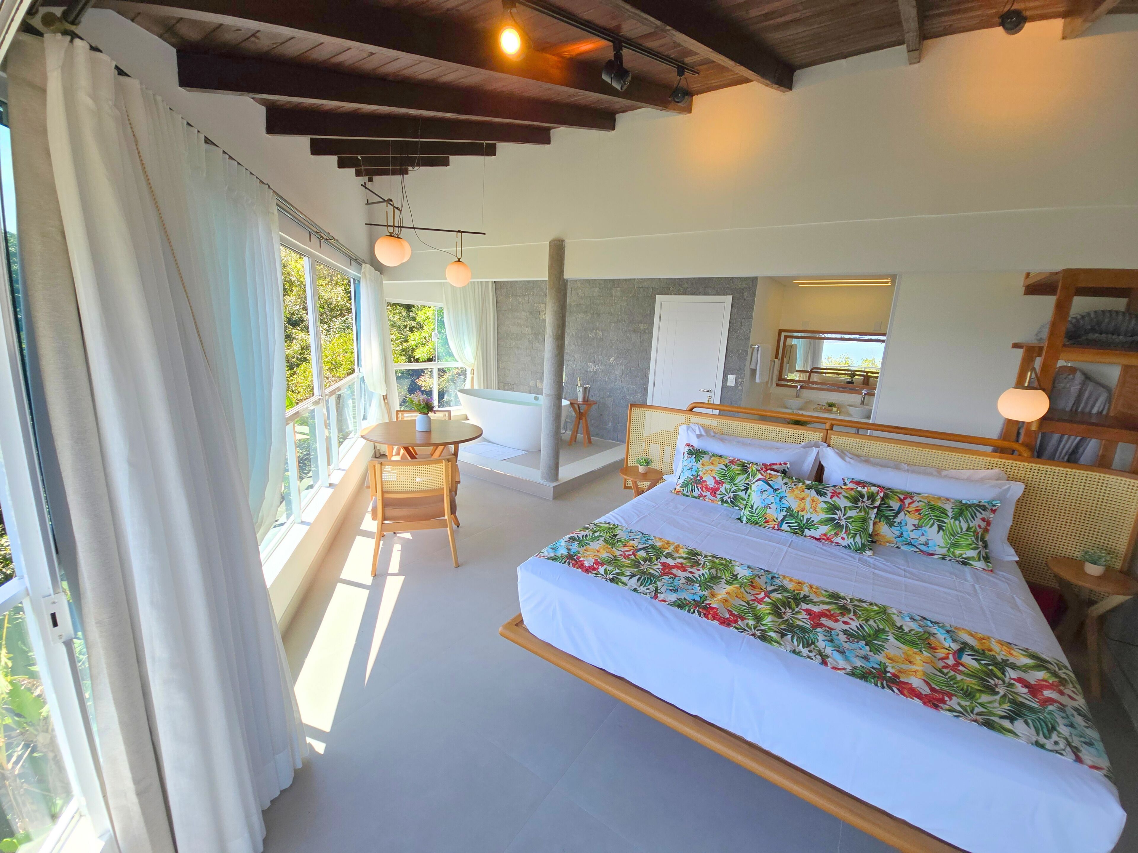 Een slaapkamer met een groot bed, een zithoek met een tafel en stoelen, een badkuip en uitzicht op groen.