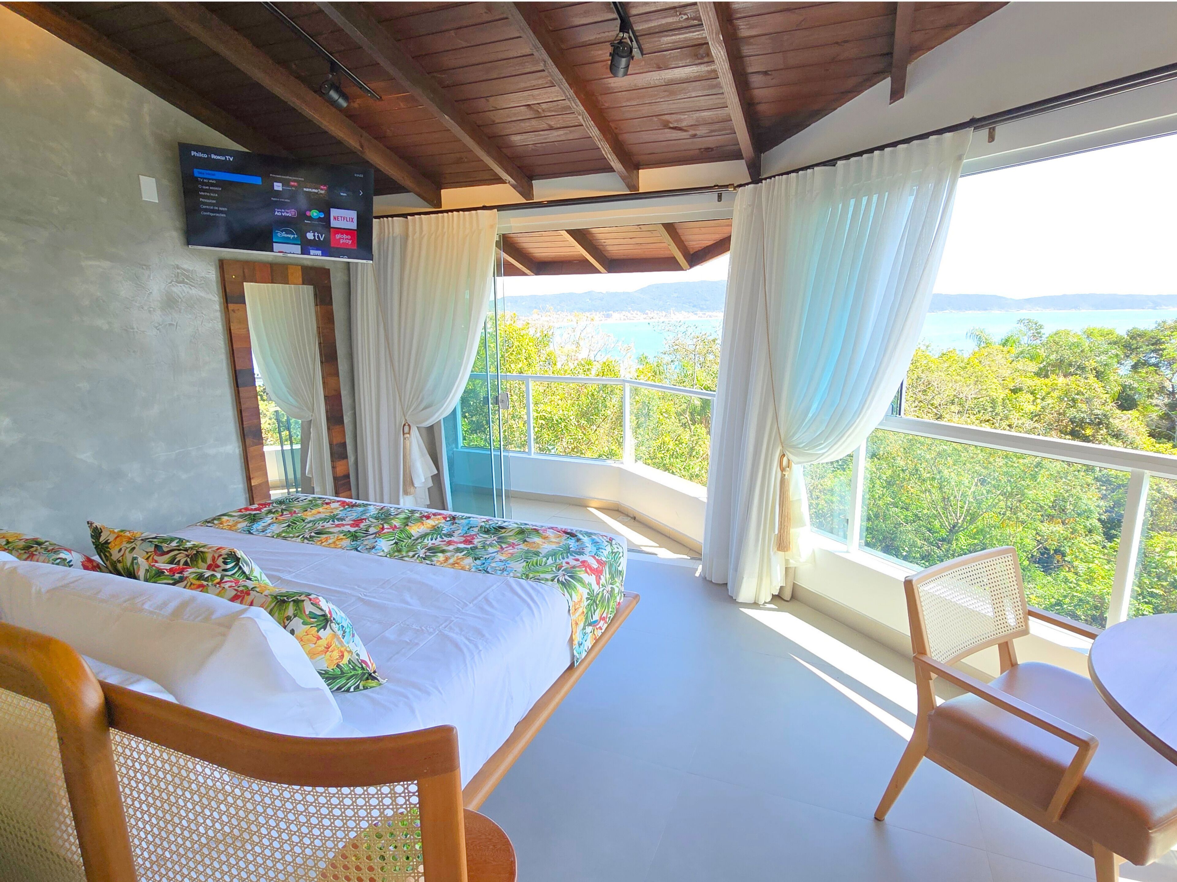 A bedroom with a bed, a chair, a TV mounted on the wall, and a balcony with a view of the landscape.