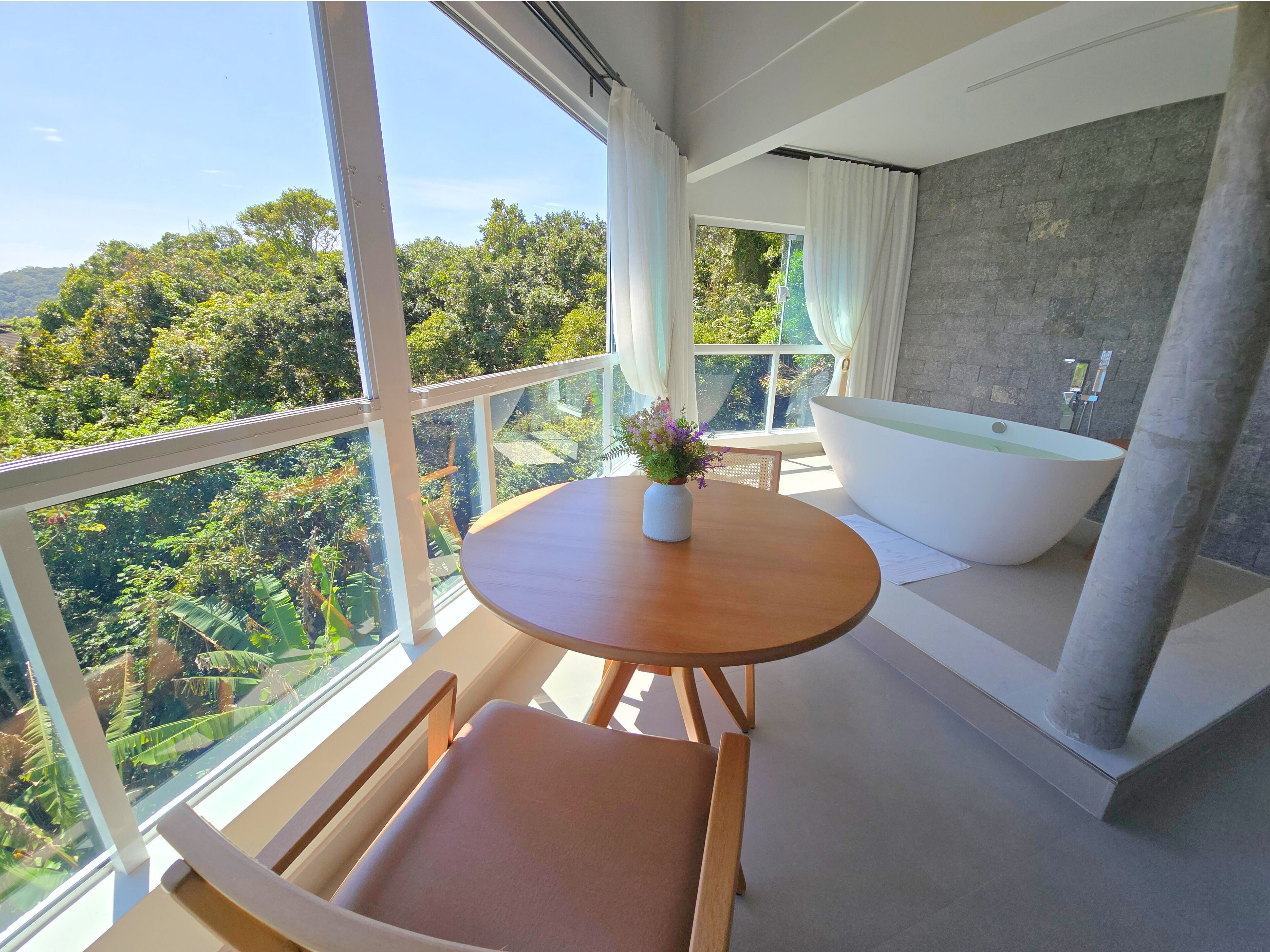 A modern bathroom with a bathtub, a round wooden table with a vase of flowers, and a view of lush greenery through large windows.