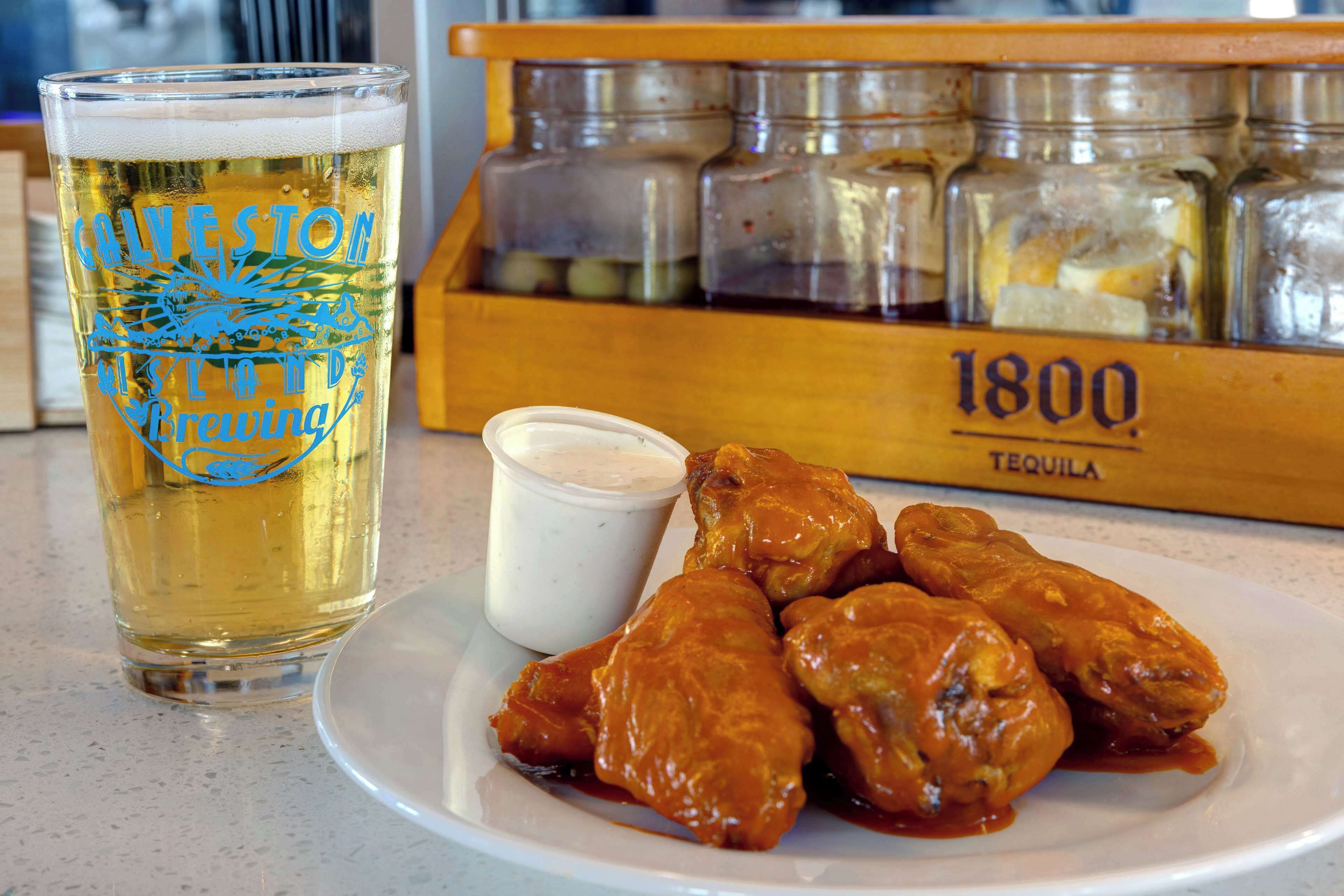 A glass of beer with a logo, a plate with wings, and a jar of sauce.