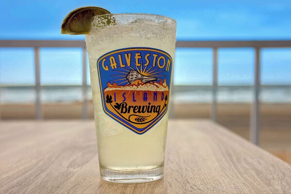 Una cerveza con una etiqueta que dice "Galveston Island Brewing" sobre una mesa de madera con una barandilla al fondo.