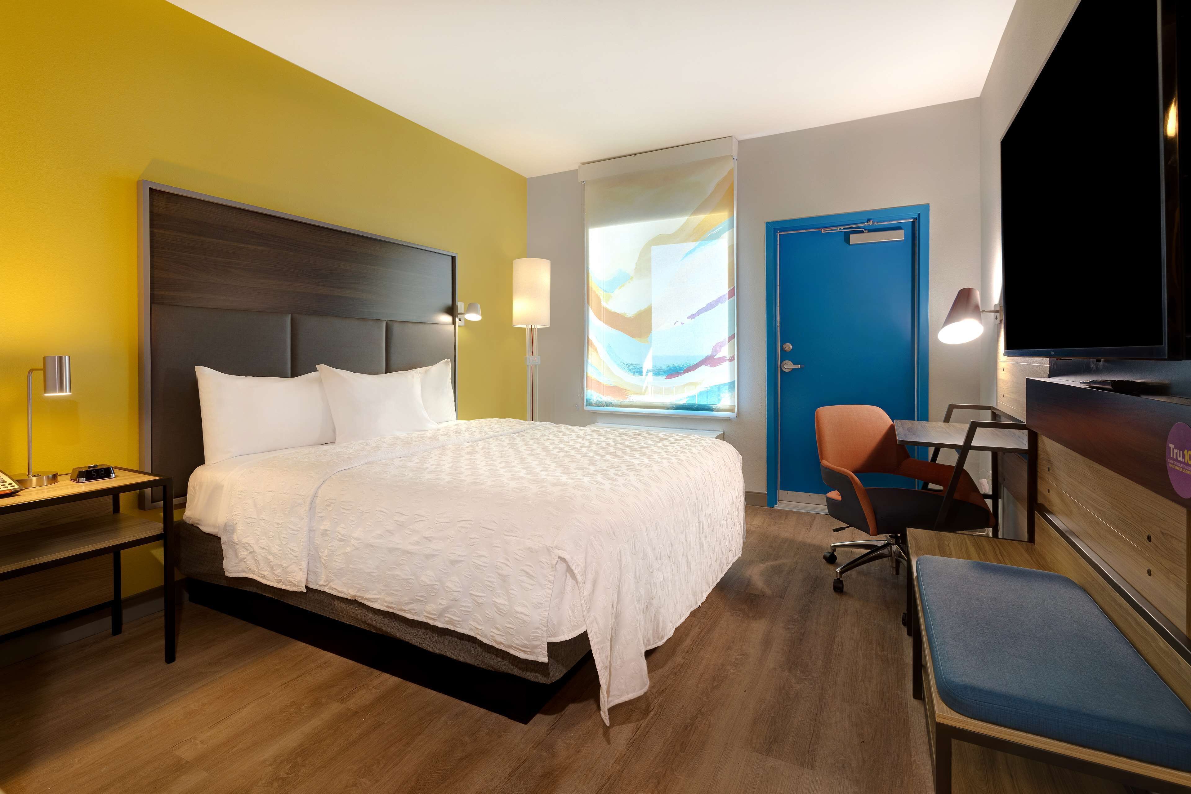 A hotel room with a large bed, a desk with a chair, a TV, and a window with a colorful view.