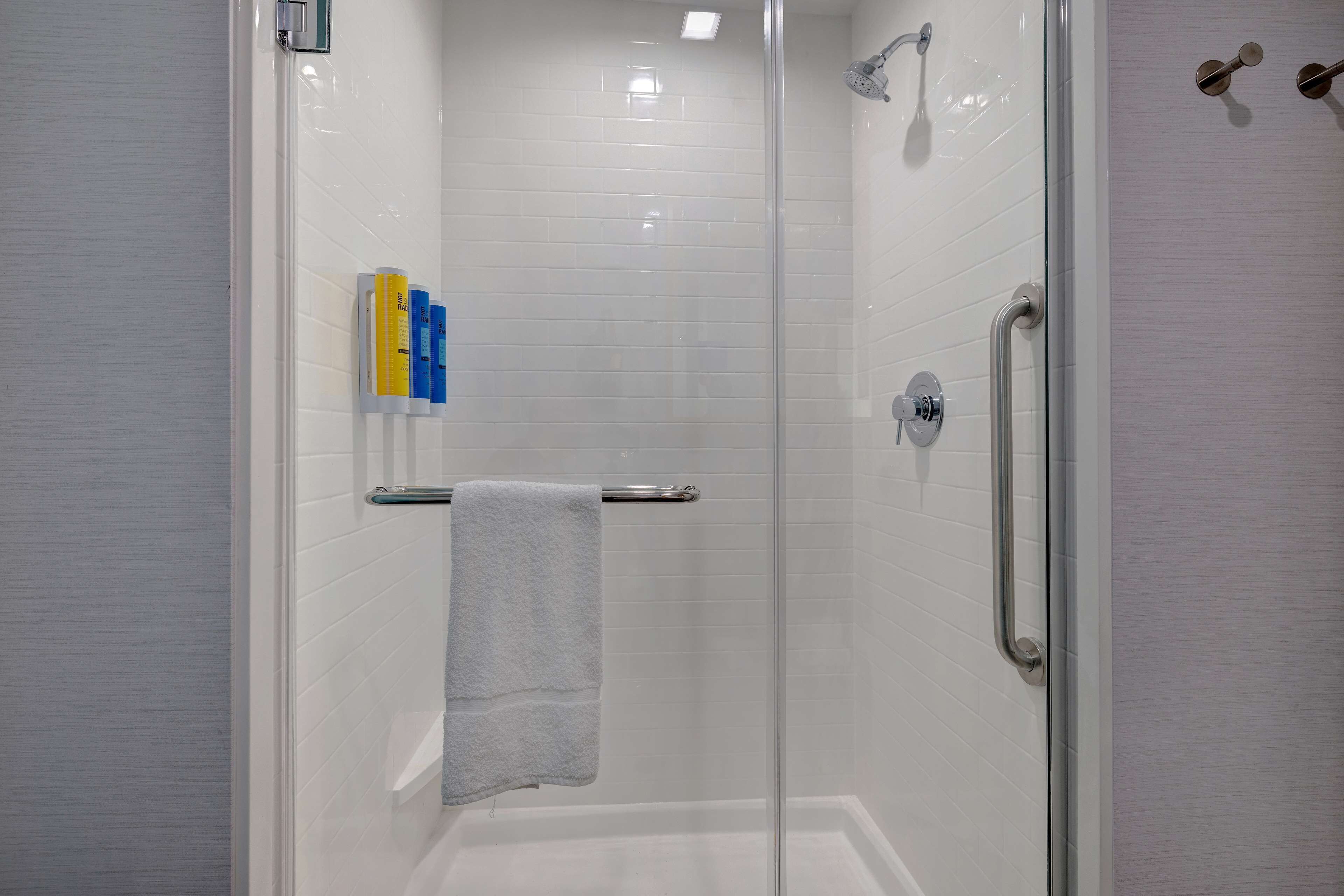 Un espace douche clos par des parois de verre, avec des murs recouverts de carreaux blancs, une porte en verre et un porte-serviettes sur lequel est posée une serviette.
