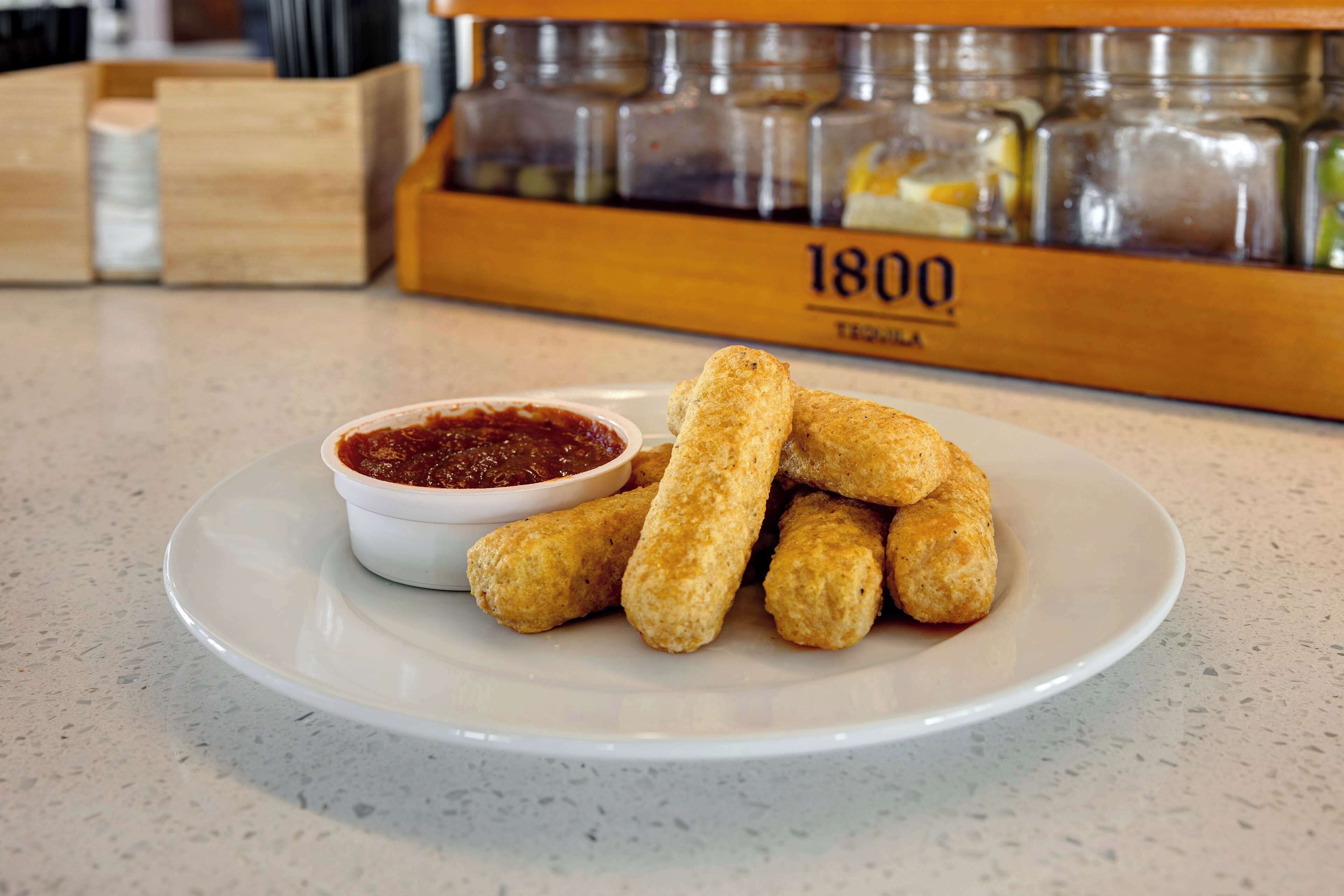 A plate of mozzarella sticks with marinara sauce on a white plate, set on a kitchen counter with a spice rack in the background.