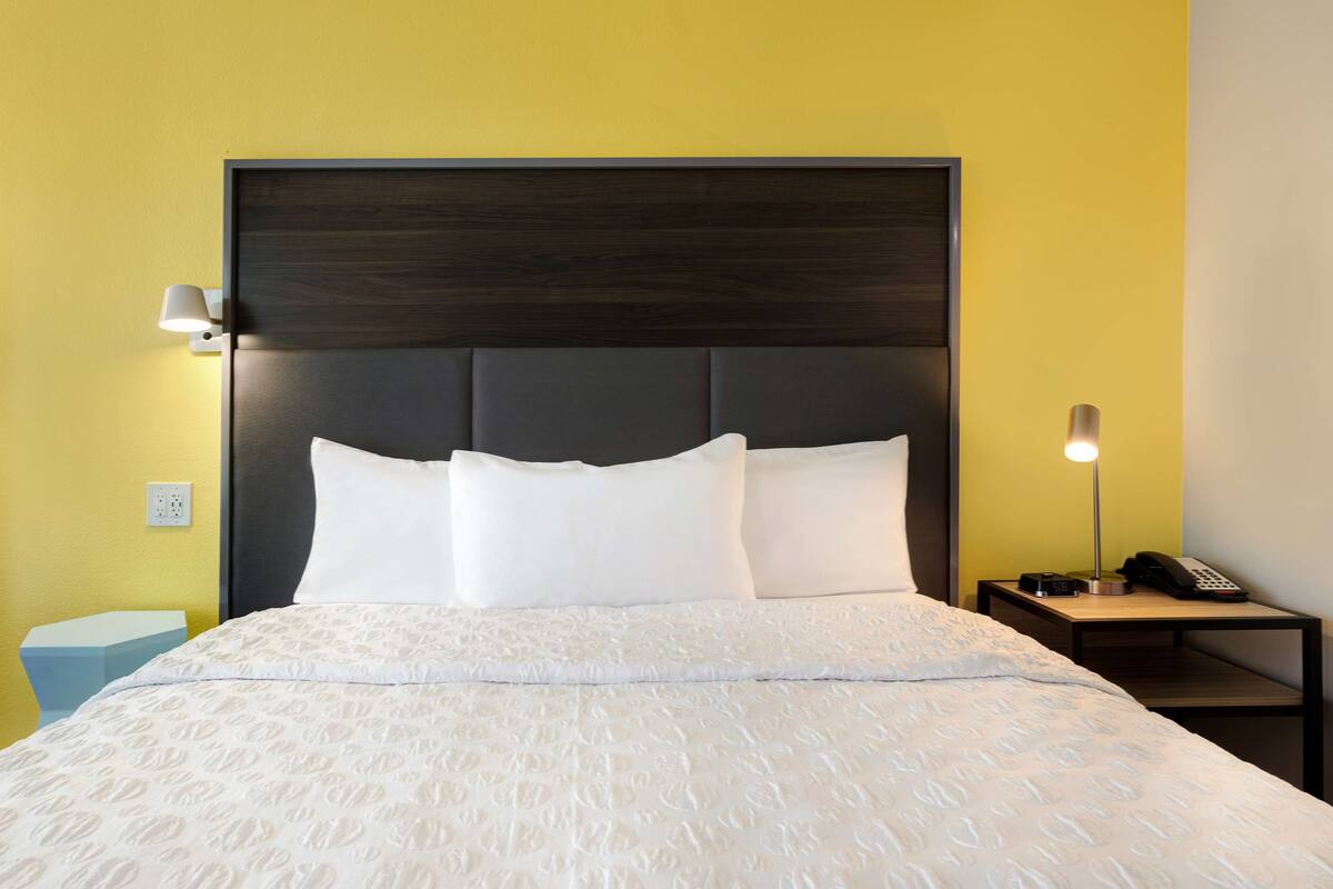Een net opgemaakt bed met witte lakens en een donker hoofdbord, een bedlampje, en een tafeltje met een telefoon en een fotocamera.