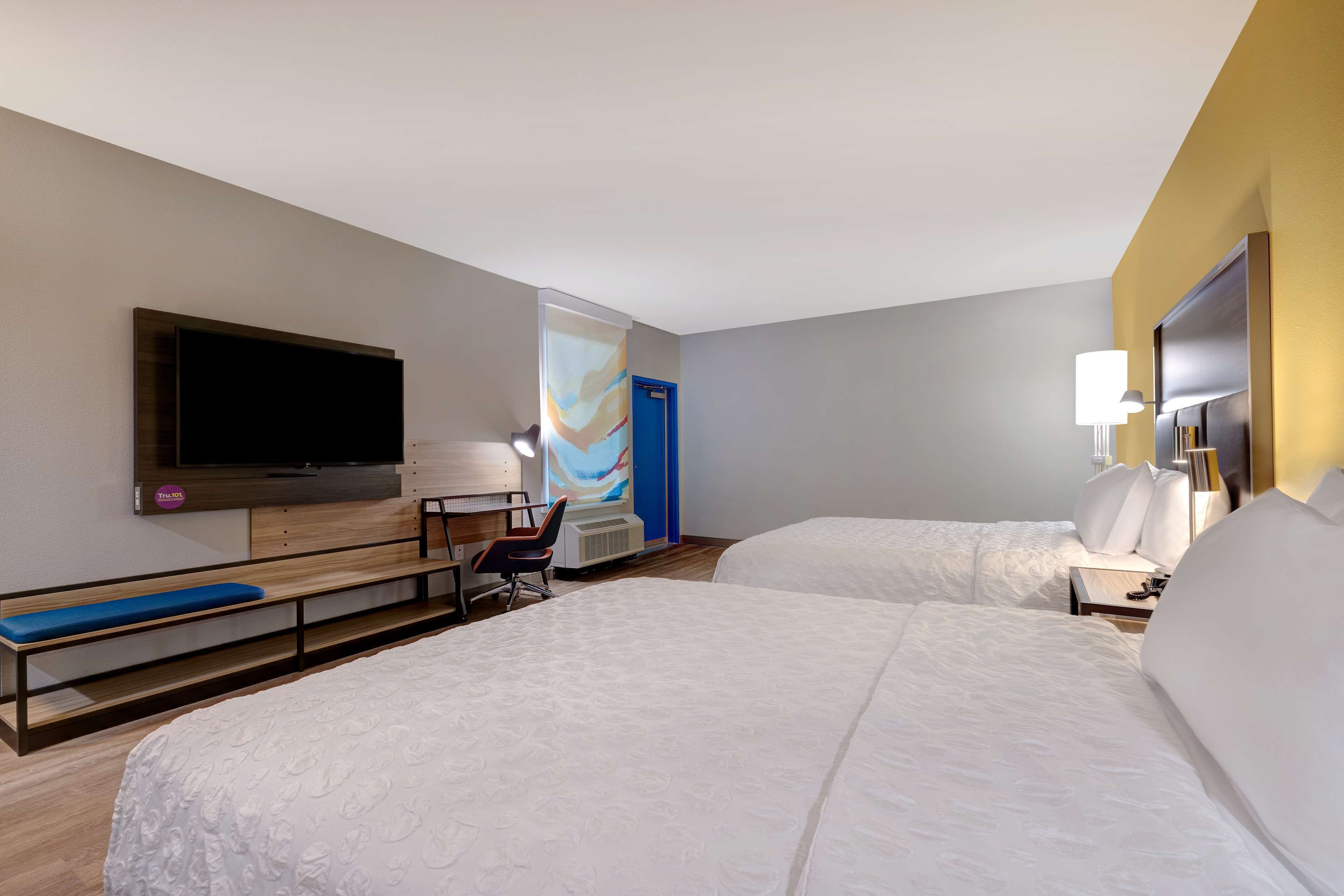 A hotel room with two beds, a flat-screen TV mounted on the wall, a desk with a chair, and a small seating area with a bench.