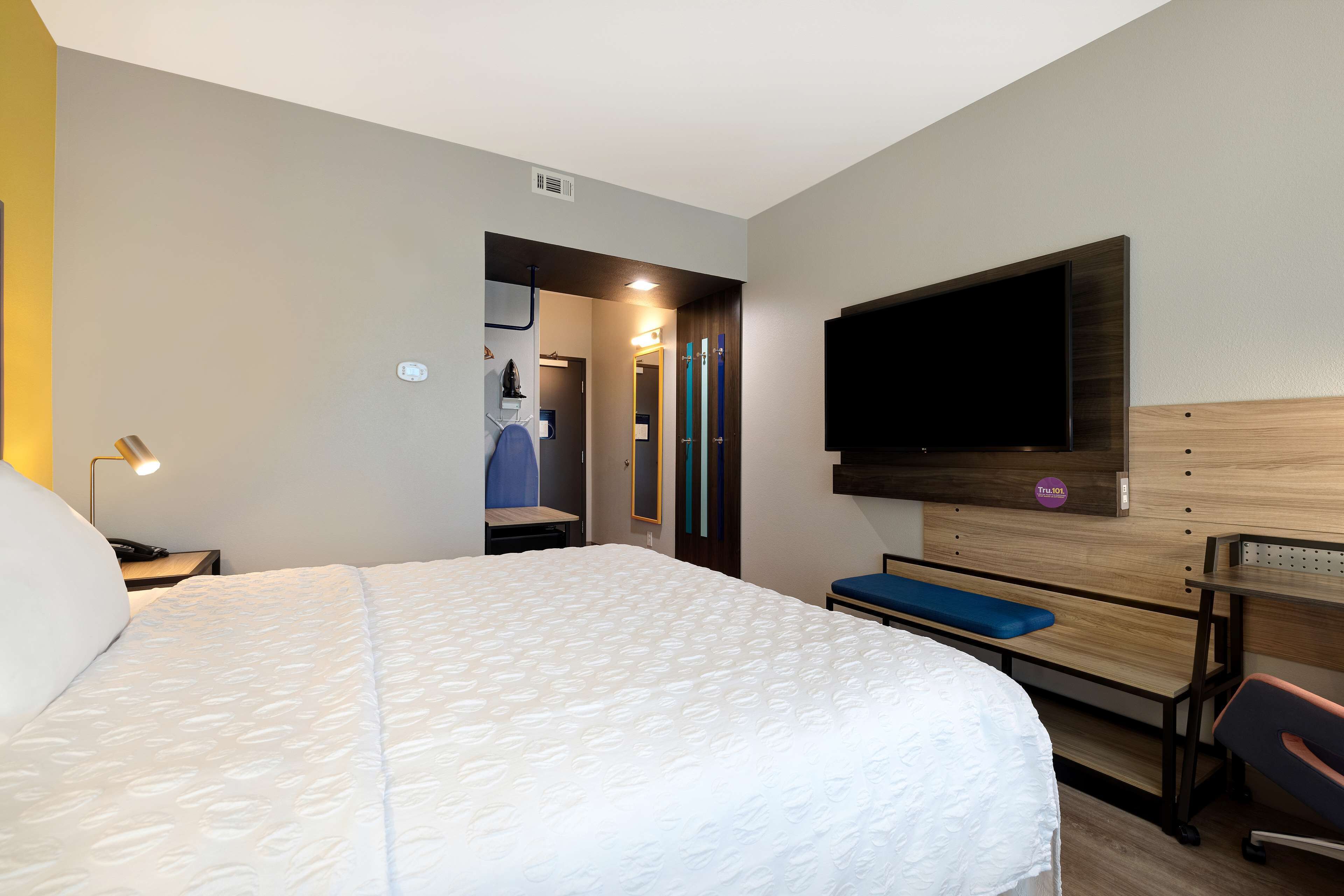 A hotel room with a large bed, a TV mounted on the wall, a desk with a chair, and a bench.