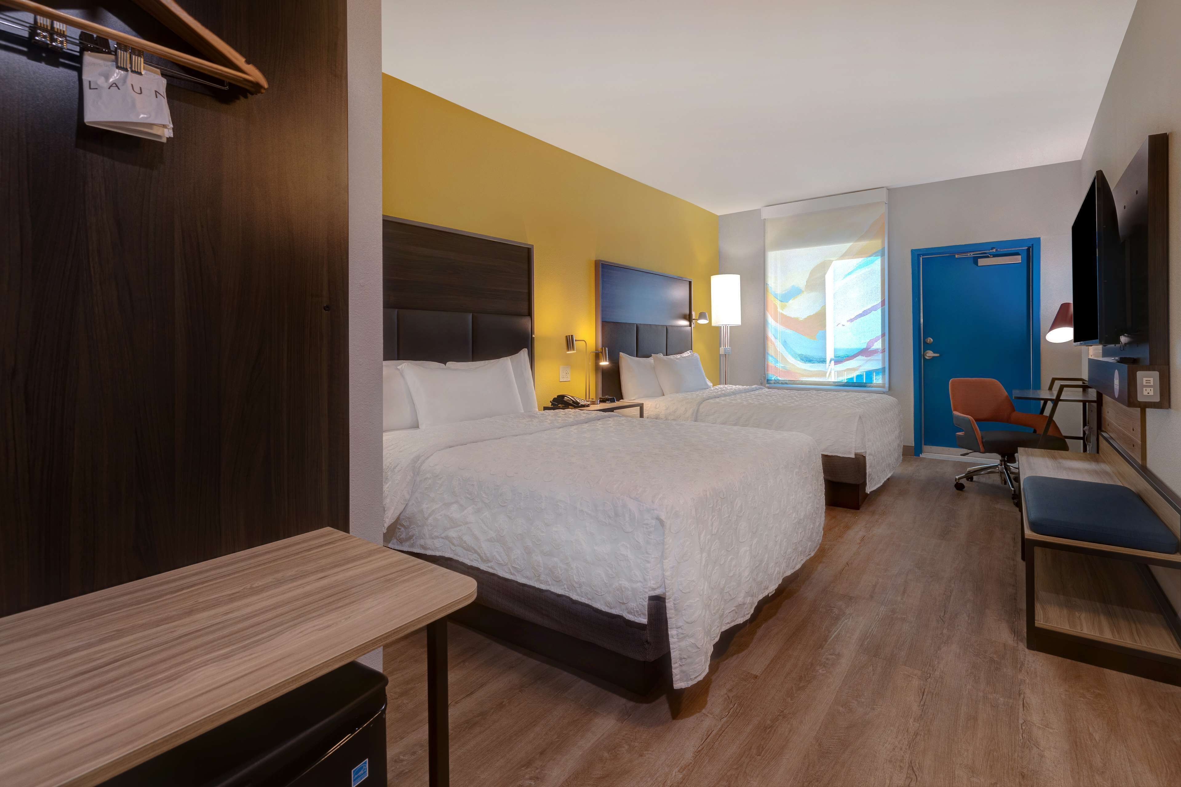 A hotel room with two beds, a desk, a chair, a TV, and a door.