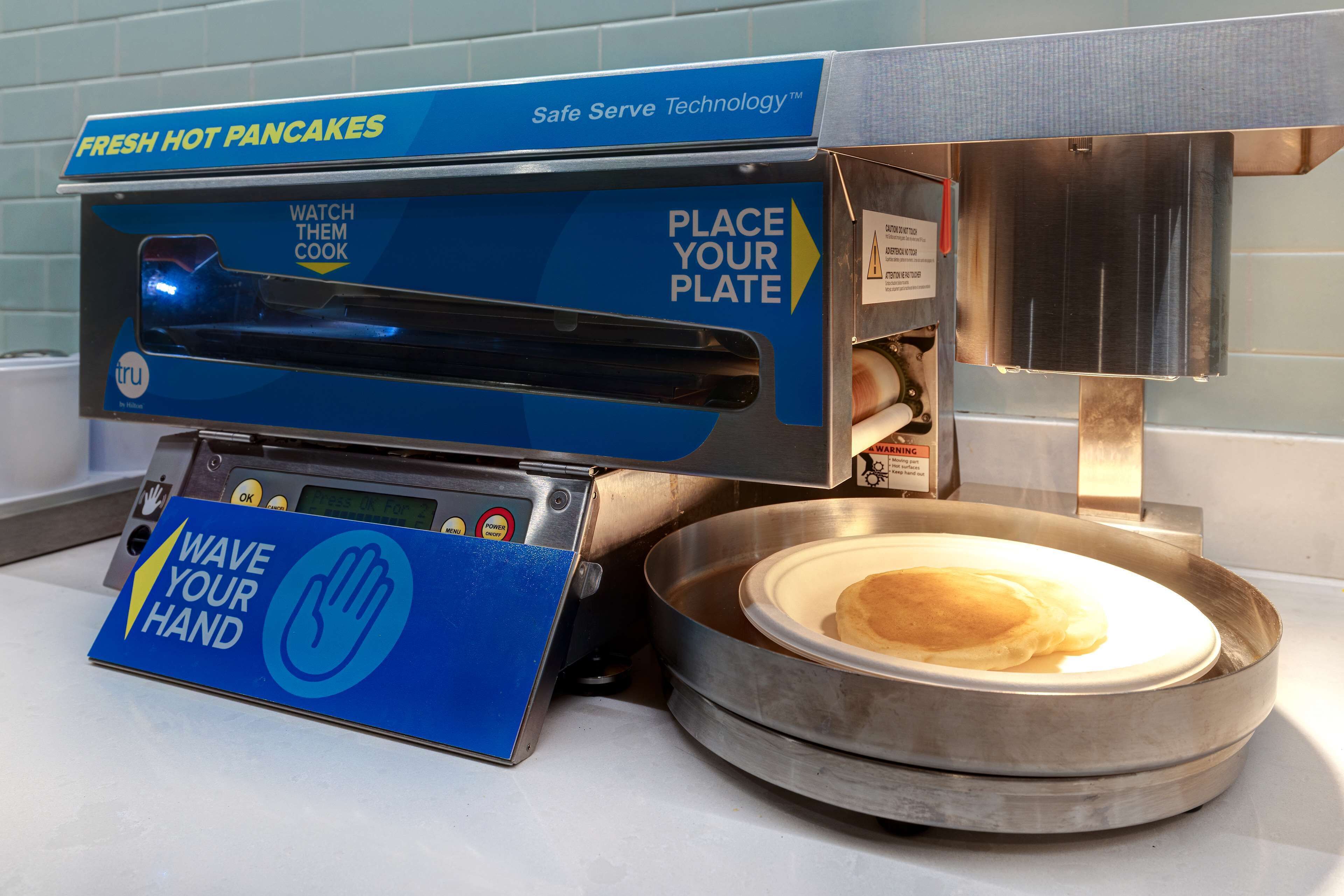A self-service pancake machine with instructions to watch them cook, place your plate, and wave your hand.