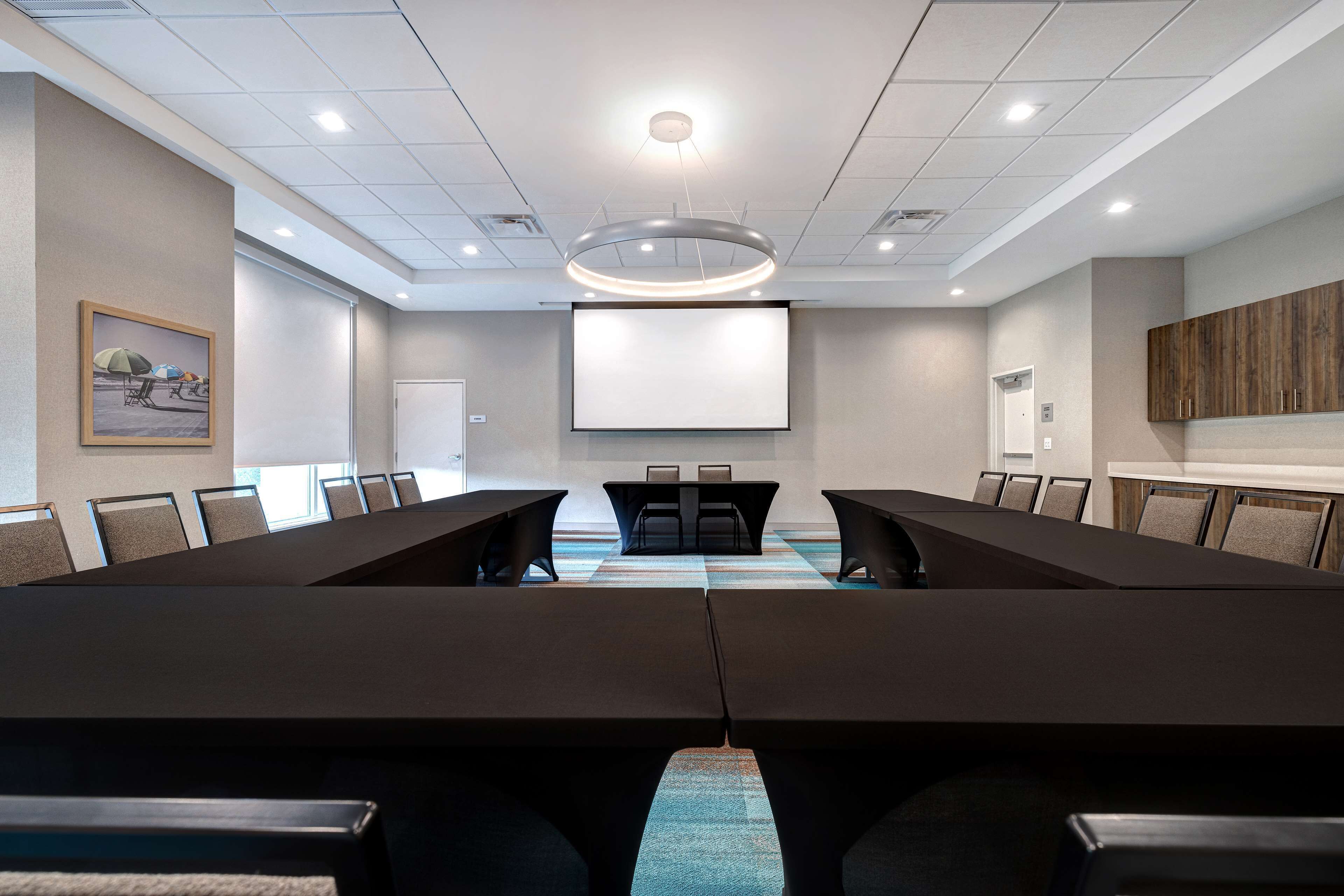 A conference room with a long table, chairs, a projector screen, and a large painting on the wall.