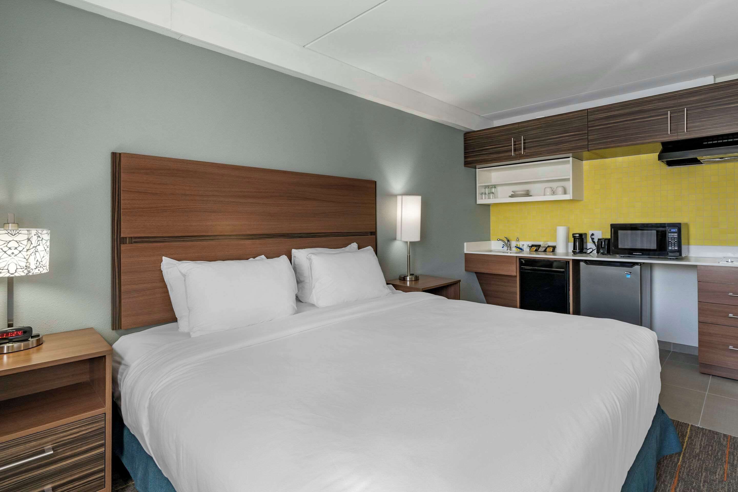 Ein Hotelzimmer mit einem großen Bett, einem Nachttisch mit Lampe, einer kleinen Küchenzeile mit Mikrowelle und einem Schrank über der Arbeitsfläche.