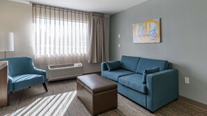 A modern living room with a blue sofa, a matching armchair, and a brown ottoman. A large window with sheer curtains allows natural light, and a painting hangs on the wall. - MainStay Suites Lexington I-75 (Lexington)
