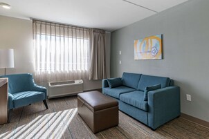 Una sala de estar moderna con un sofá azul, un sillón a juego y un reposapiés marrón. Un ventanal con cortinas translúcidas deja entrar luz natural, y un cuadro adorna la pared.