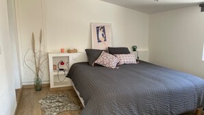 Un dormitorio con una cama grande, una mesita de noche, una lámpara, una obra de arte en la pared y una planta decorativa.