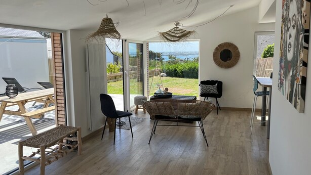 Amplia sala de estar con un gran ventanal corredero que da a un patio, un sofá de mimbre, una silla negra y una mesa de comedor con sillas.