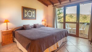 Une chambre dotée d’un grand lit, de deux tables de chevet avec des lampes, d’une œuvre d’art encadrée au mur et d’une porte-fenêtre coulissante donnant sur un espace extérieur aménagé avec des chaises.