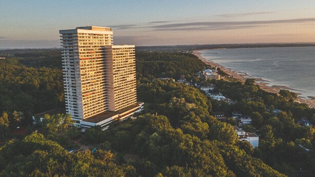 Ein Hochhaus-Hotel mit mehreren Stockwerken, umgeben von üppiger Vegetation und einem Strand im Hintergrund.