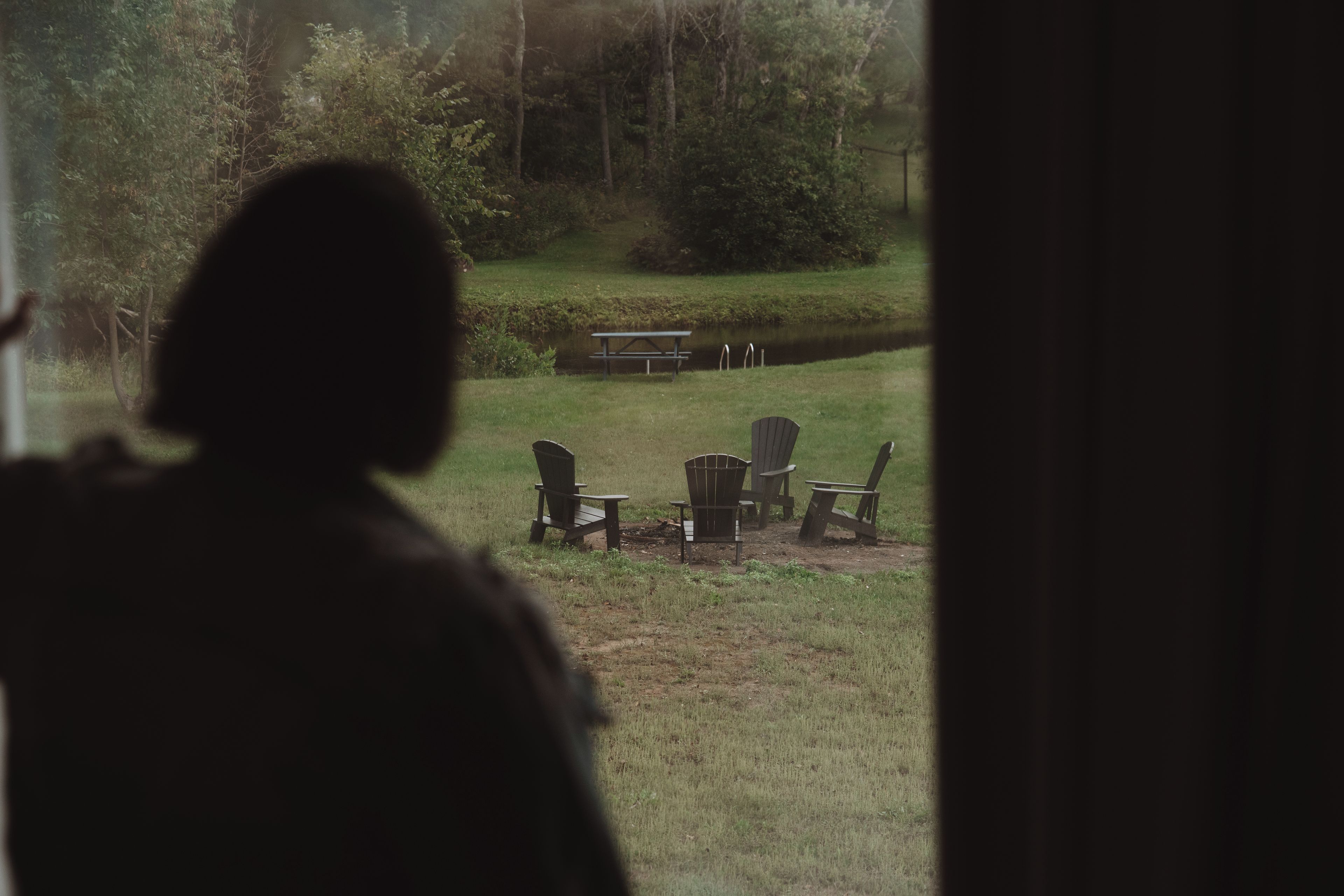 A silhouette of a person standing by a window overlooking a grassy area with outdoor seating.