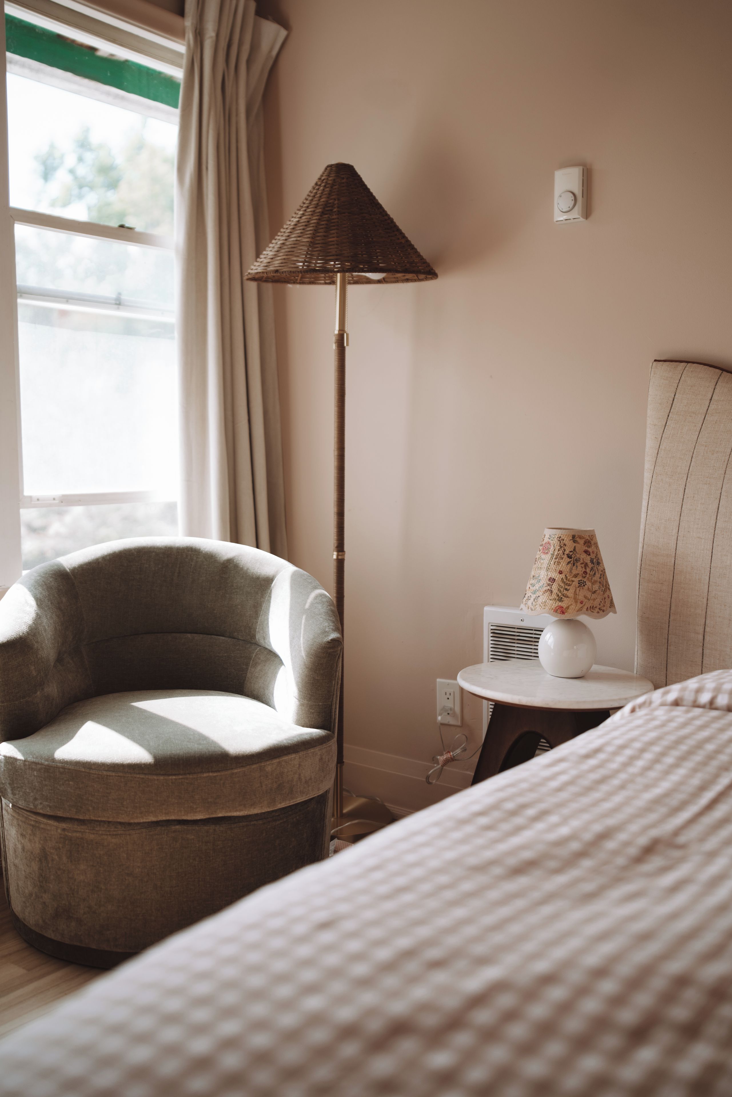 A hotel room with a round armchair, a floor lamp, a bedside table with a lamp, and a bed.