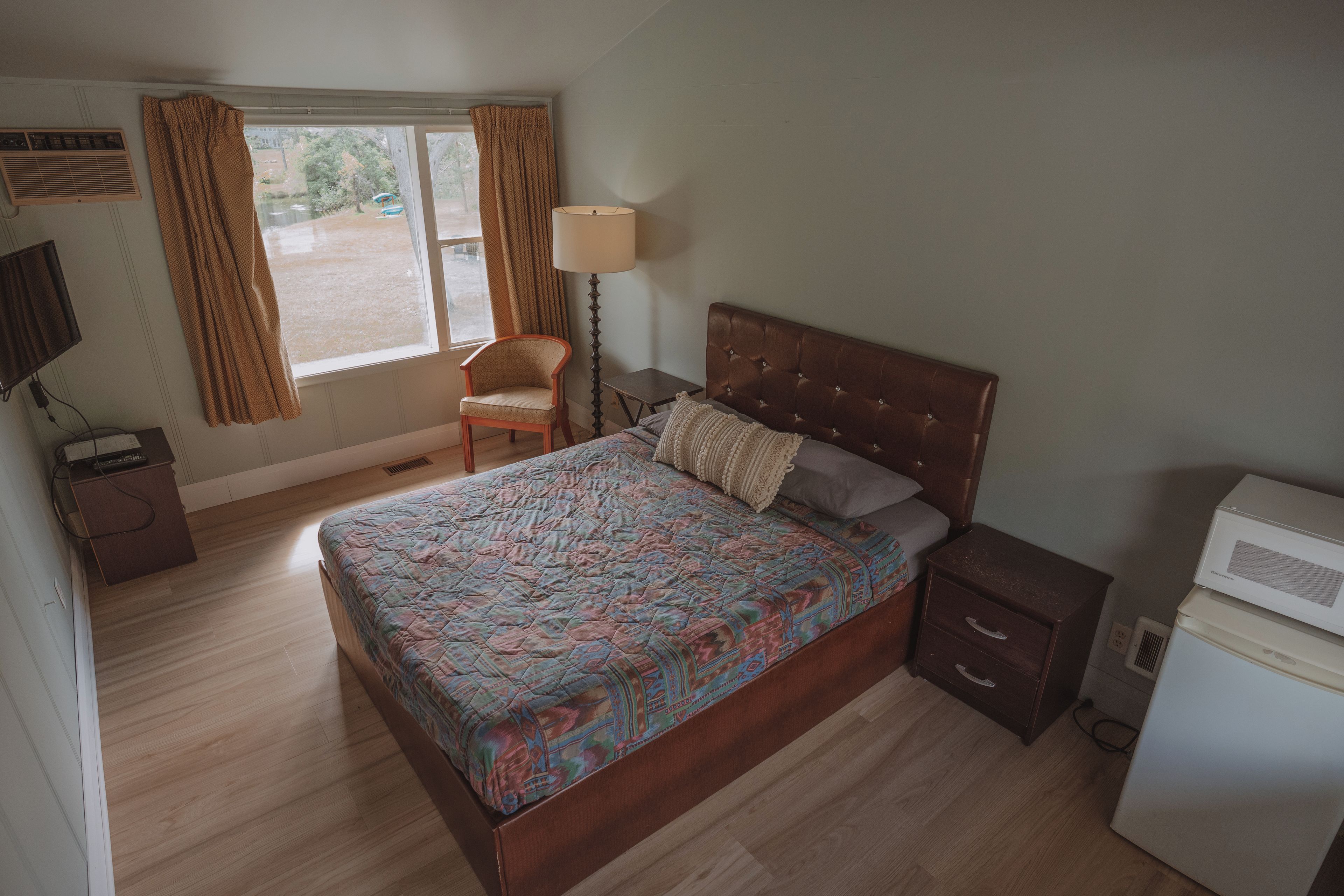 Un dormitorio con cama, una silla, una mesita de noche, una lámpara y una ventana con cortinas.