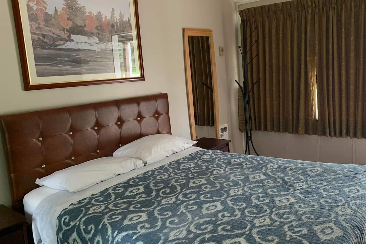 Un dormitorio con una cama grande, cabecera, un cuadro enmarcado en la pared, una ventana con cortinas y una puerta.